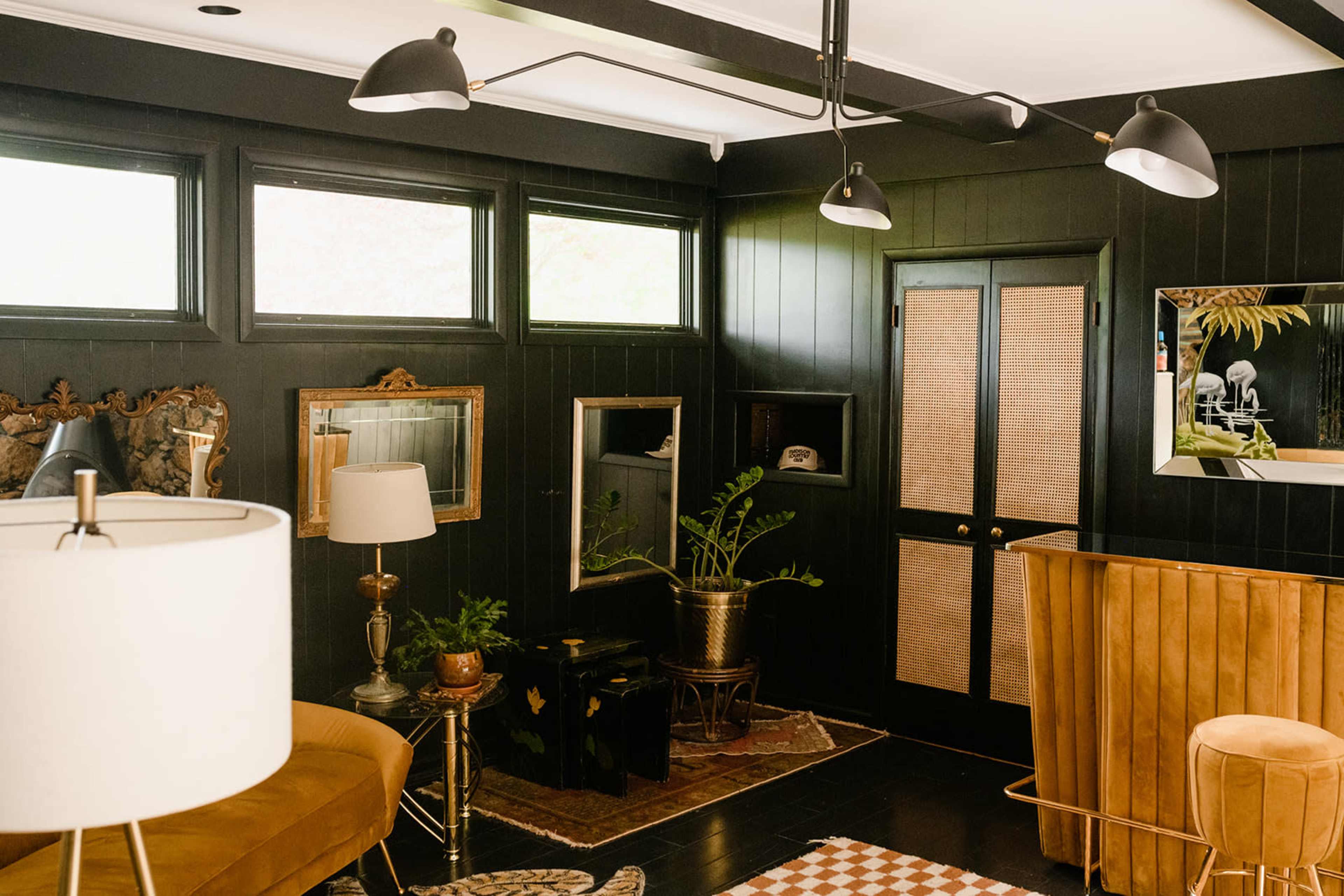A dark-colored room featuring a blend of vintage and modern furnishings, including a lamp, a mid-century chair, and a wooden bar.