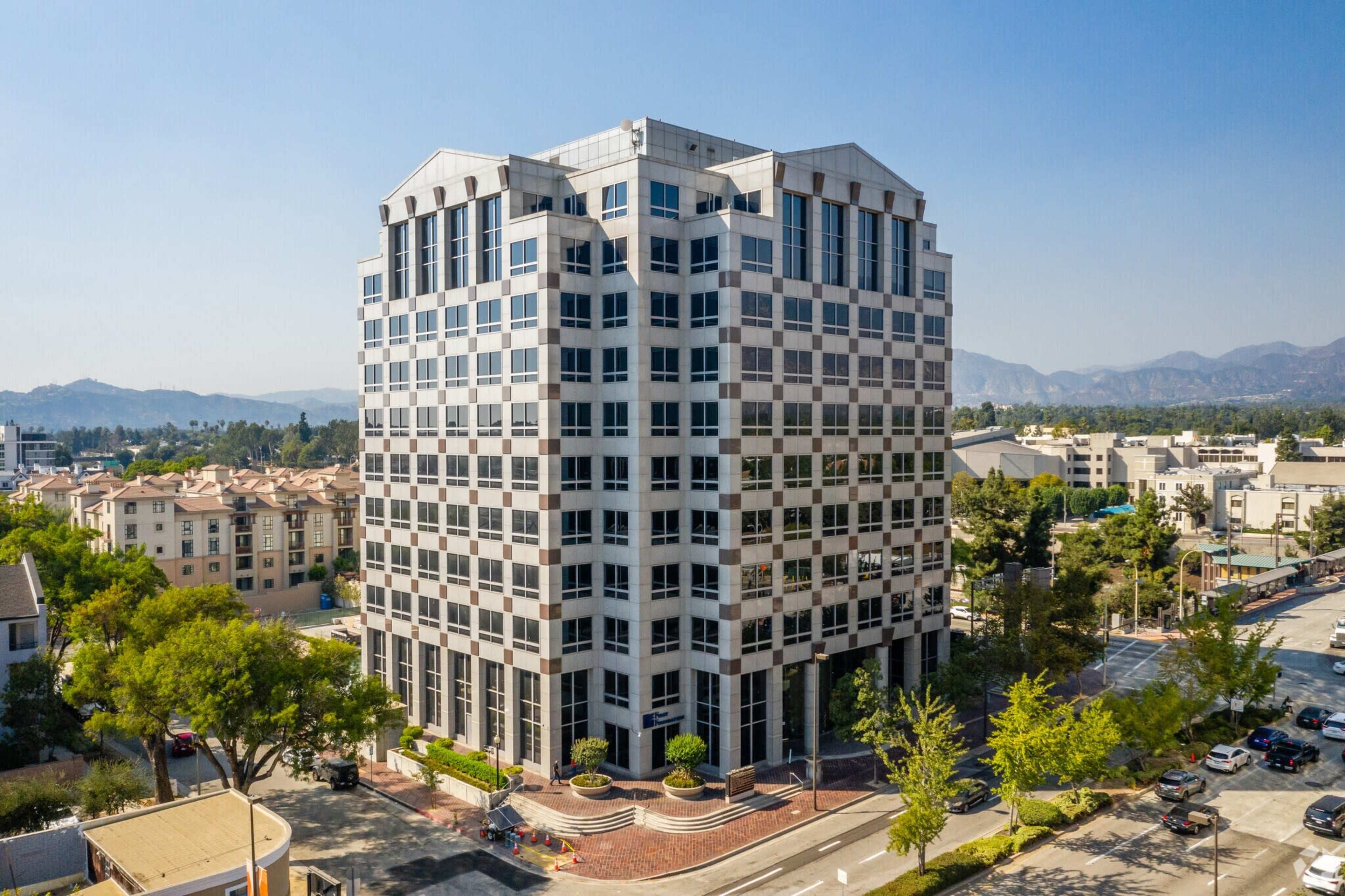 A modern, multi-story office building with large windows stands prominently at the corner of a city street, surrounded by trees and residential units.