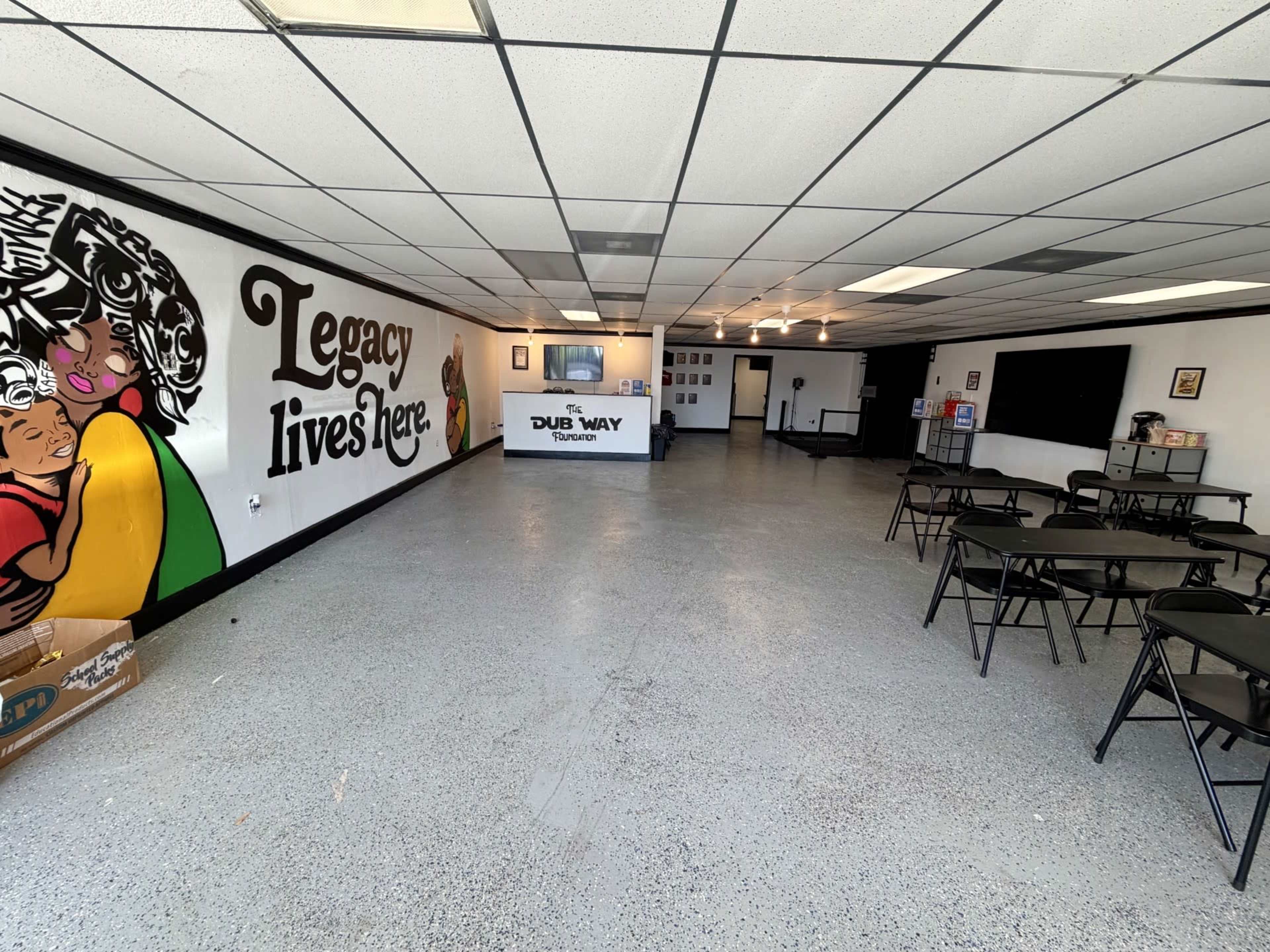 The image shows a spacious, empty indoor area with a mural featuring the words "Legacy lives here" on one wall and several tables set up throughout the room.