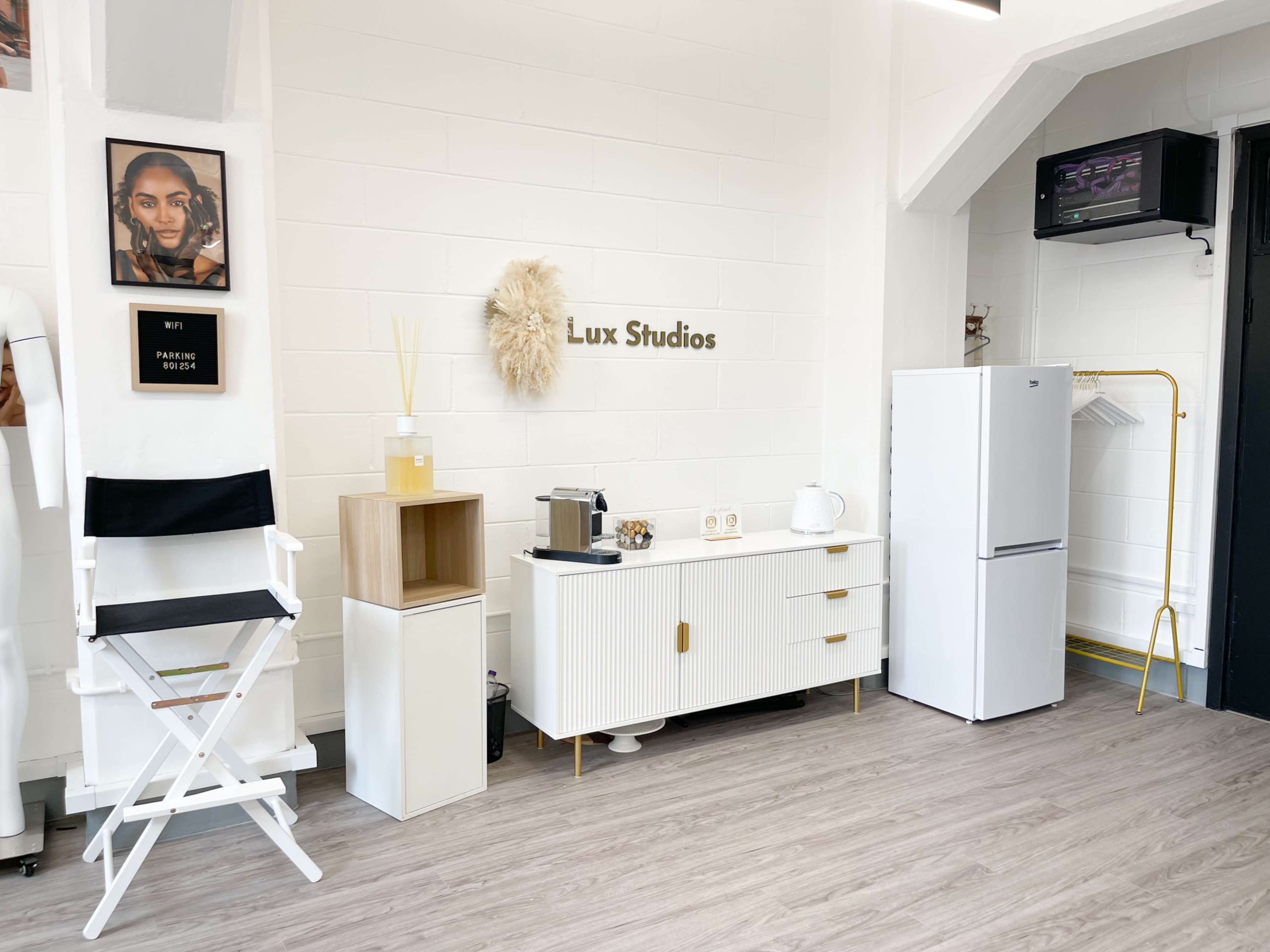 A modern studio interior featuring a white cabinet, a refrigerator, a coffee station, a director's chair, and artwork on the walls.