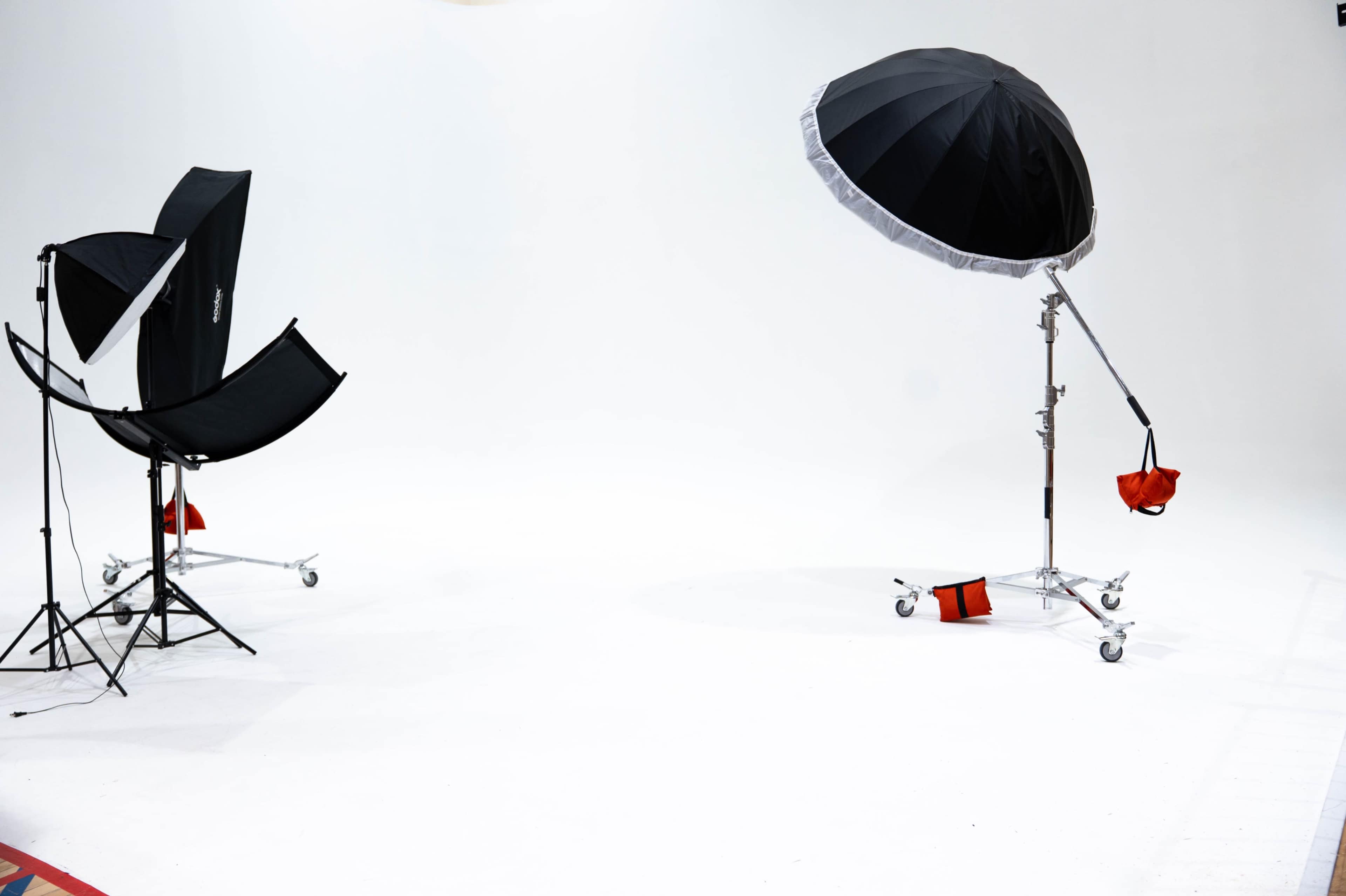The image shows a photography studio with a white backdrop, featuring a softbox on a stand and an umbrella light on a wheeled support, accompanied by several small red bags on the floor.