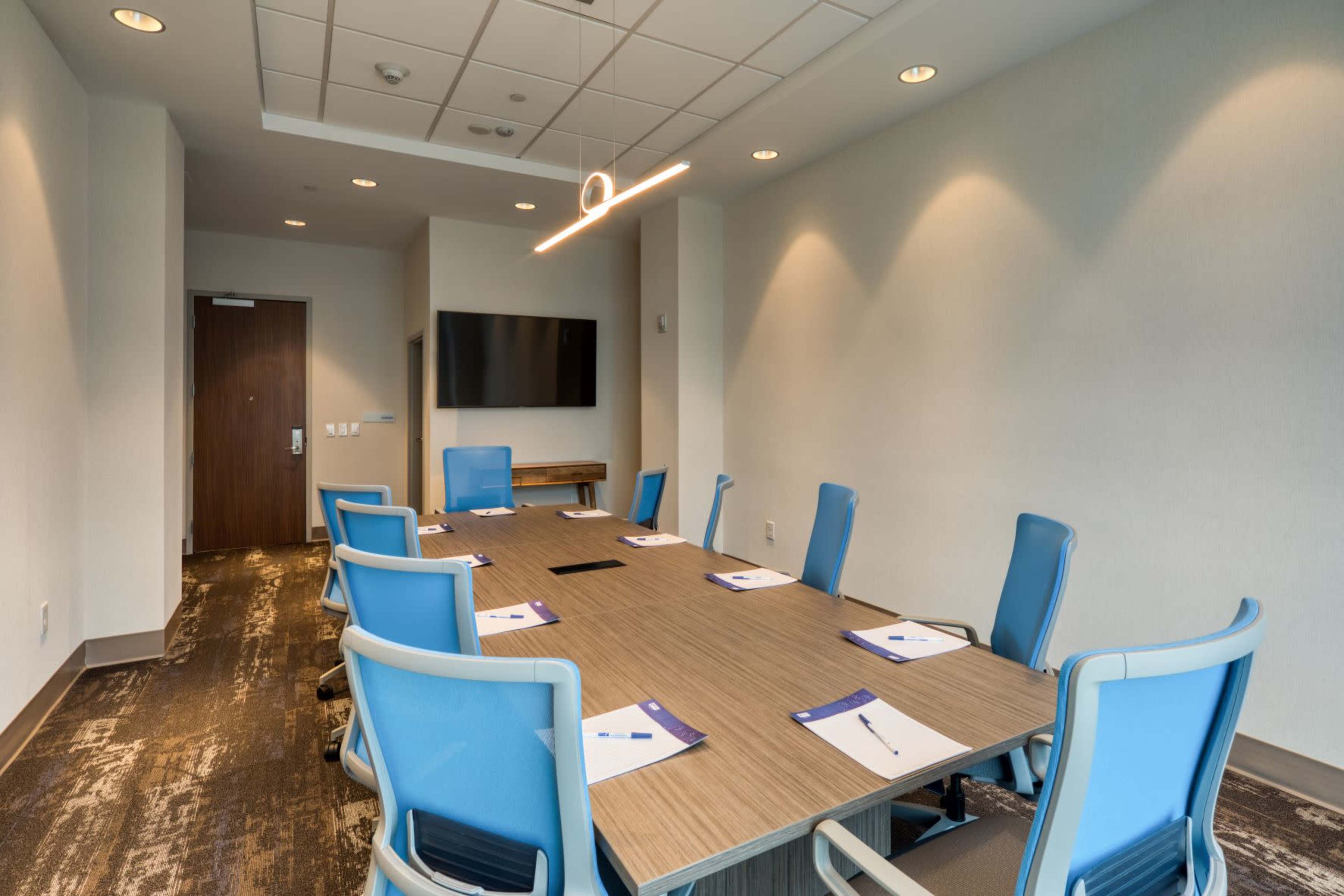 The image shows a modern conference room with a long wooden table, surrounded by blue chairs, and featuring a wall-mounted television and notepads on the table.