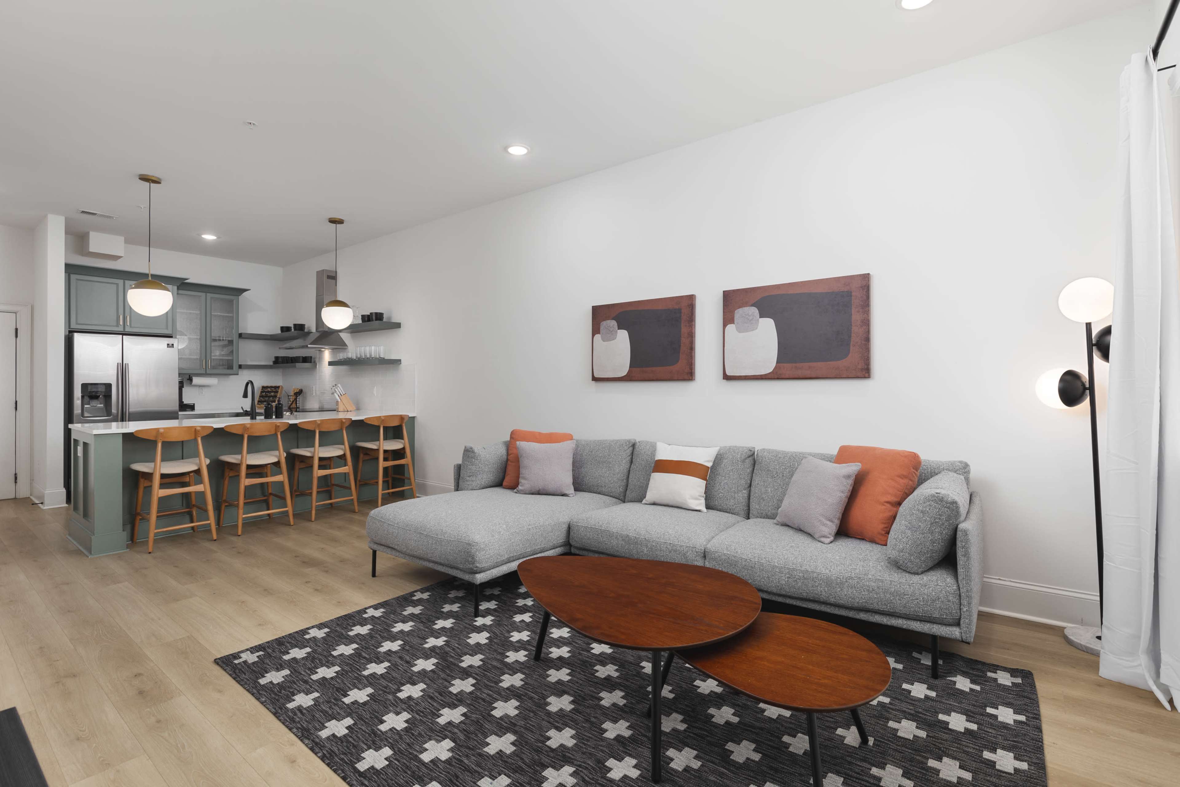 A modern living room with a gray sectional sofa, two wooden coffee tables, and a kitchen area featuring bar stools and green cabinetry.