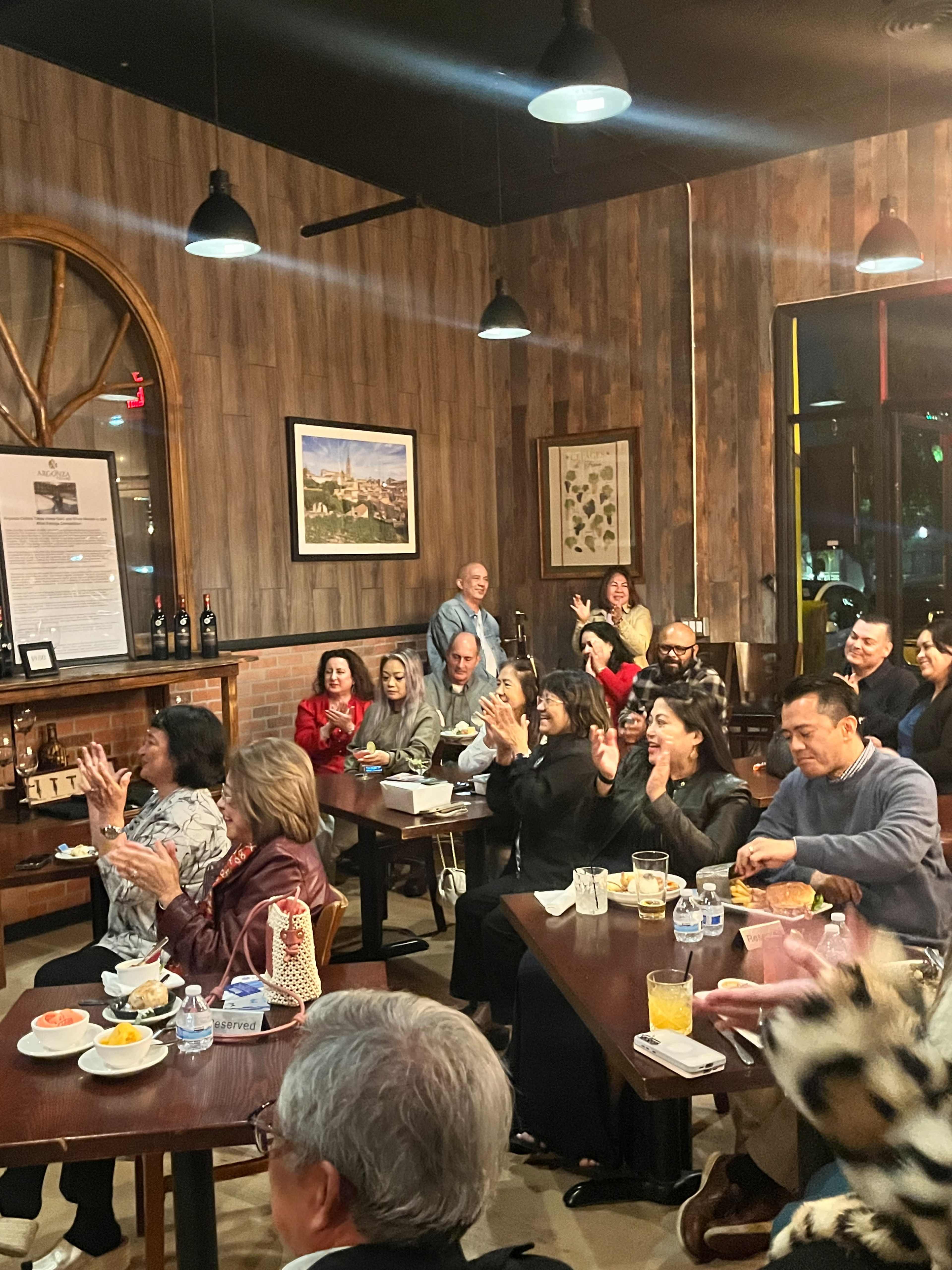 A diverse group of people is gathered inside a restaurant, clapping and enjoying food and drinks while sitting at tables.