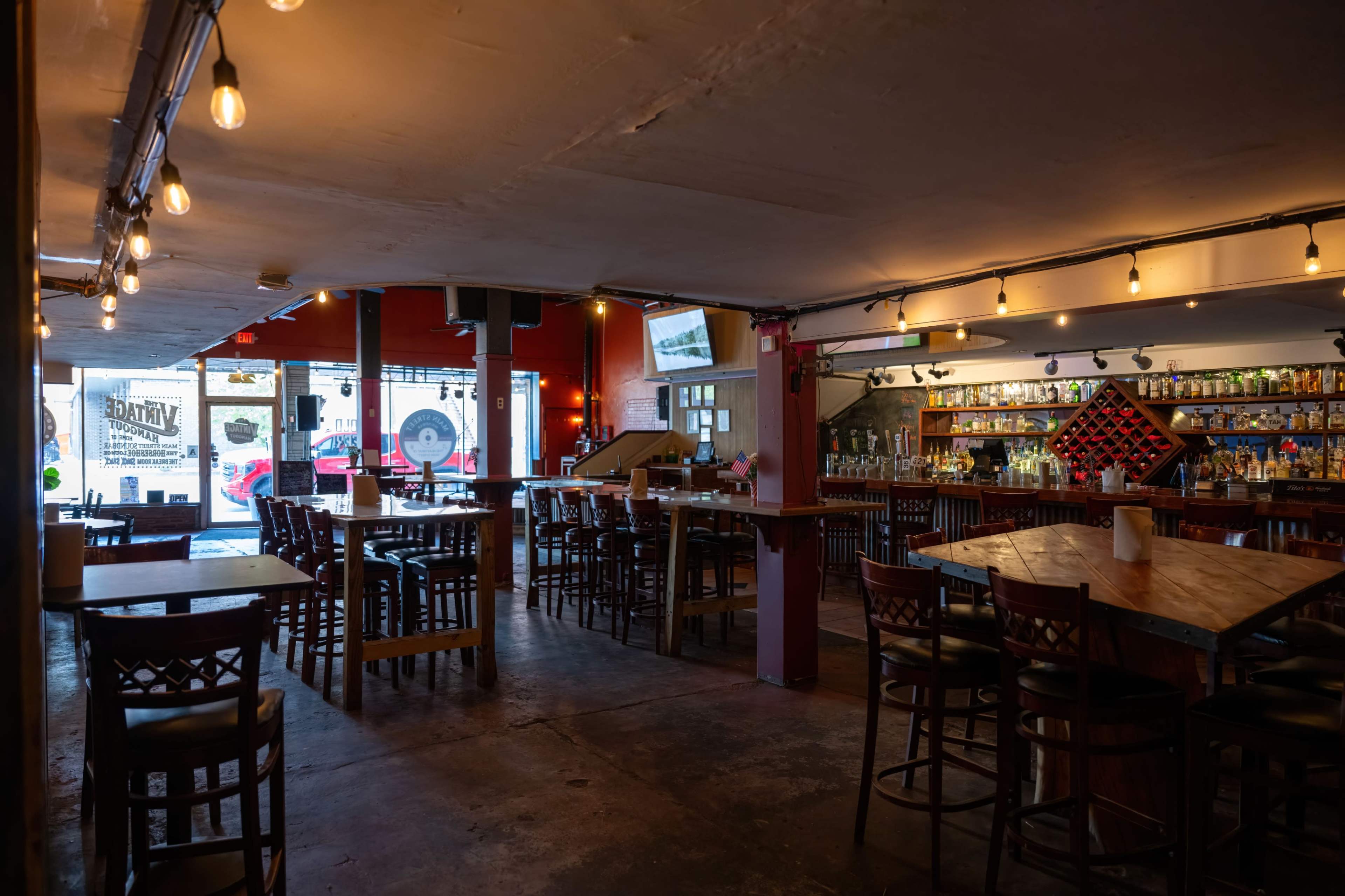 The image shows an empty bar interior with wooden tables and chairs, warm lighting, and a wall lined with shelves of liquor bottles.