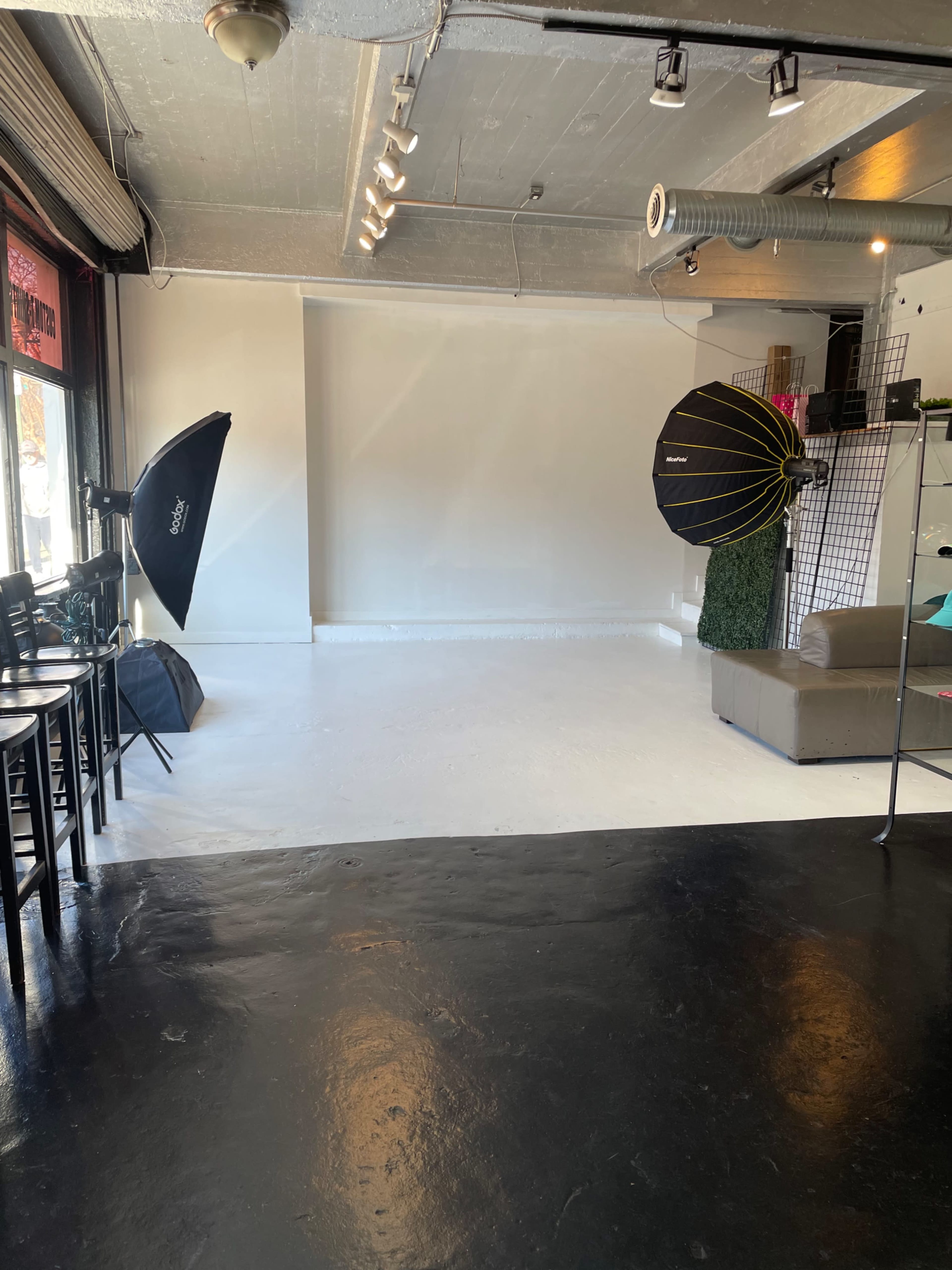 A spacious studio with a blank white backdrop, two softbox lights, and a seating area featuring a sofa and chairs.