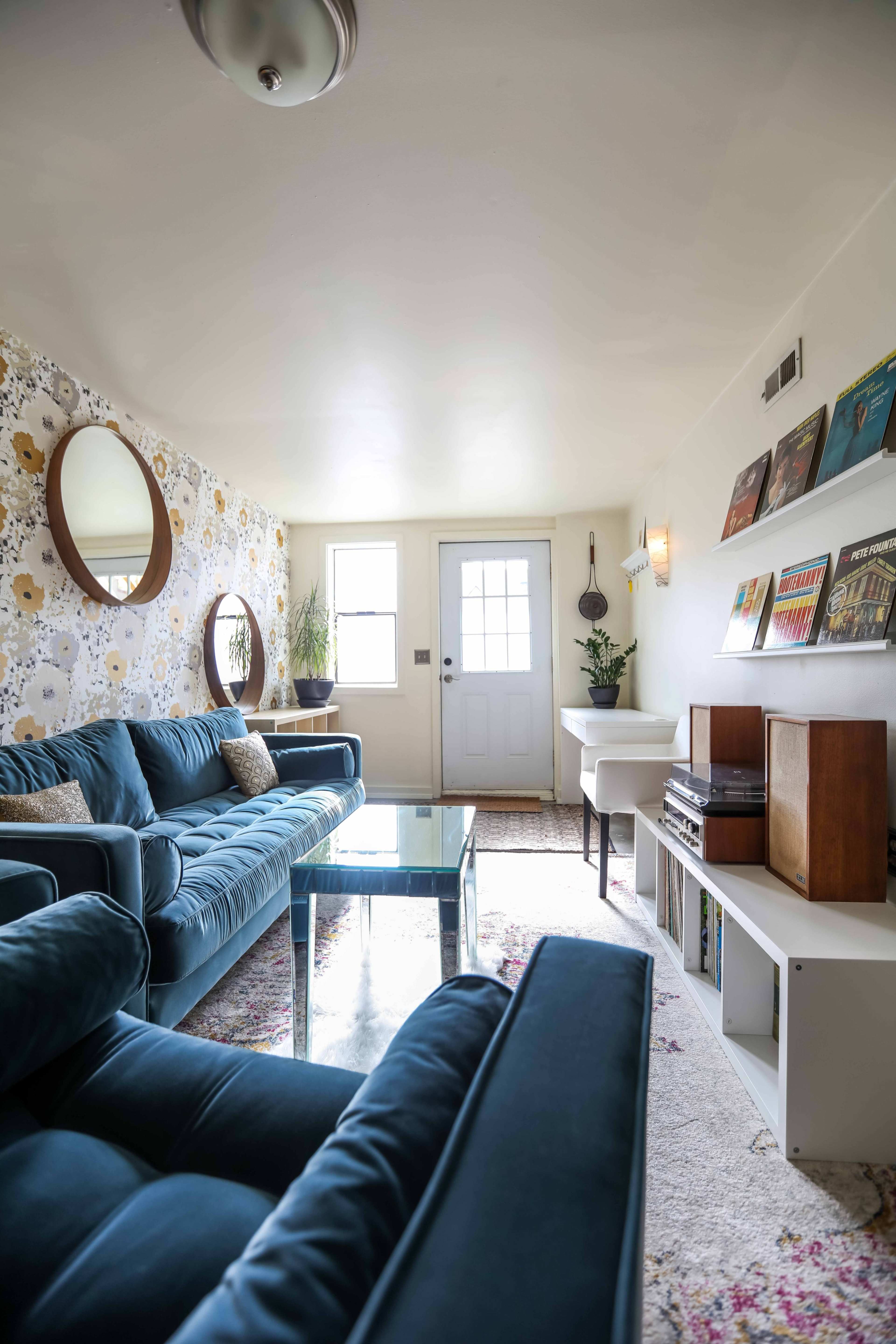 The image shows a living room with blue sofas, a glass coffee table, a door leading outside, and shelves displaying records and books.
