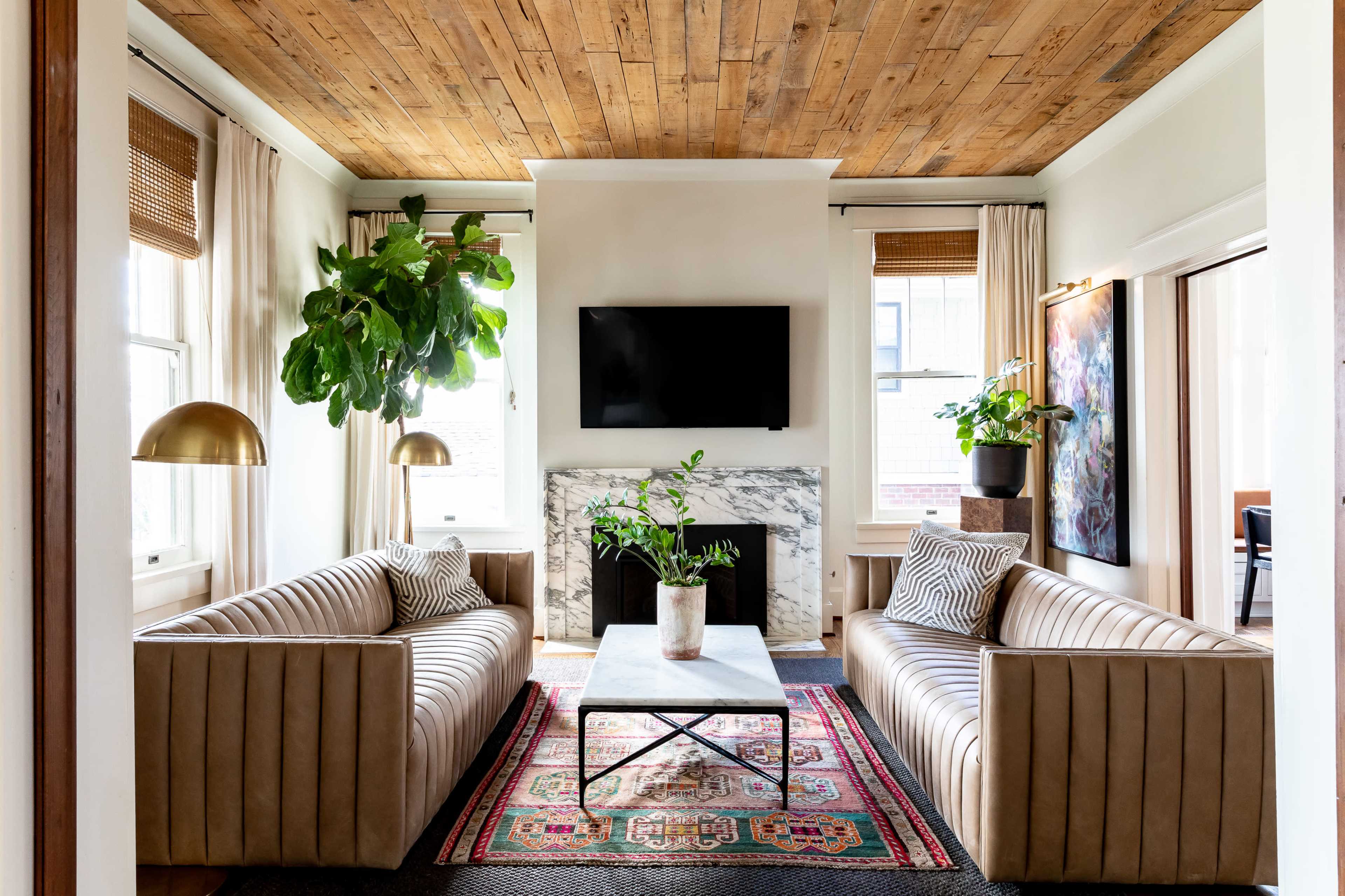 The image showcases a cozy living room featuring two beige sofas, a coffee table with a plant, a marble fireplace, and a mounted television on a wooden ceiling backdrop.