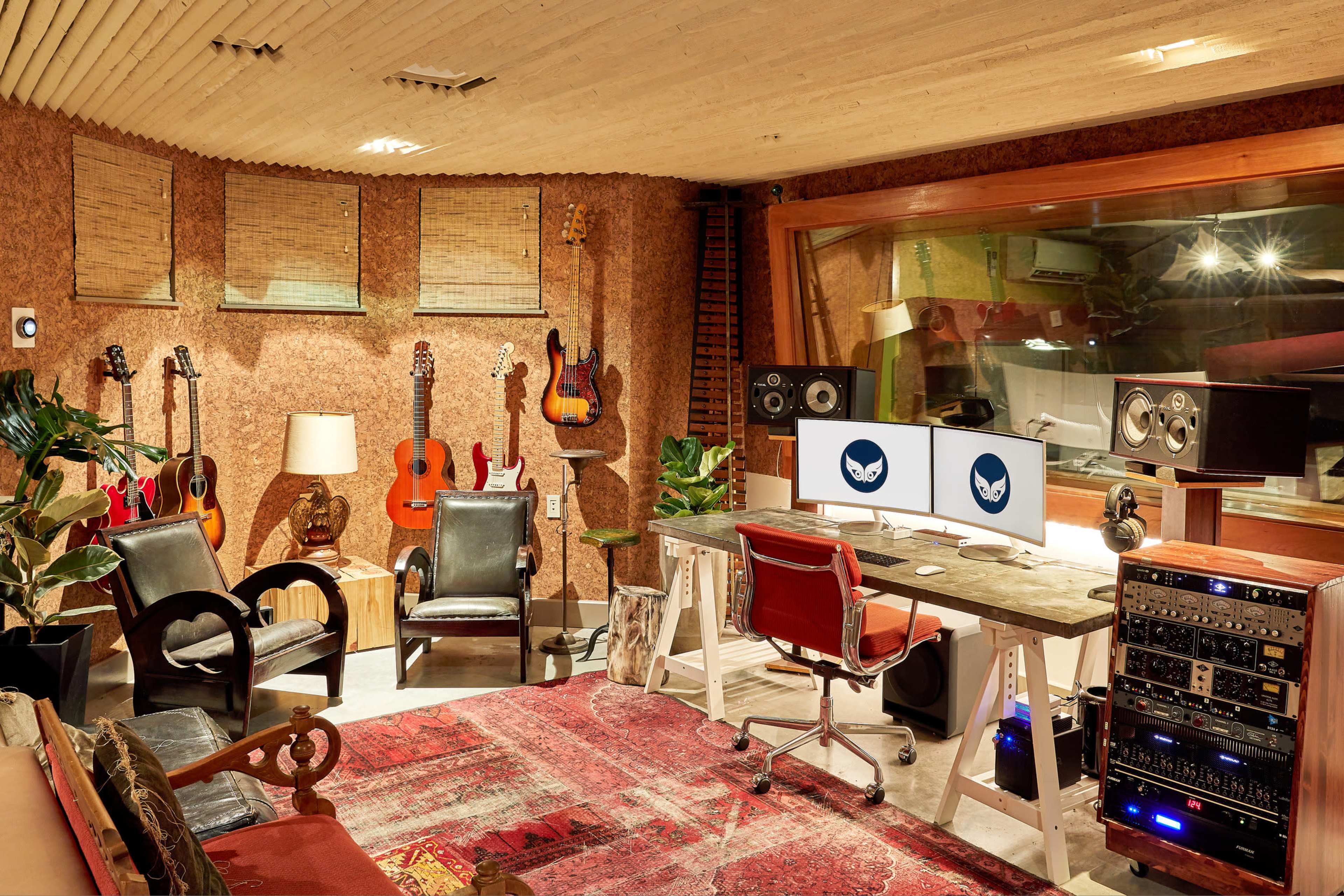 A music studio with a desk featuring dual monitors, audio equipment, and various guitars displayed on the walls, surrounded by seating and decorative elements.
