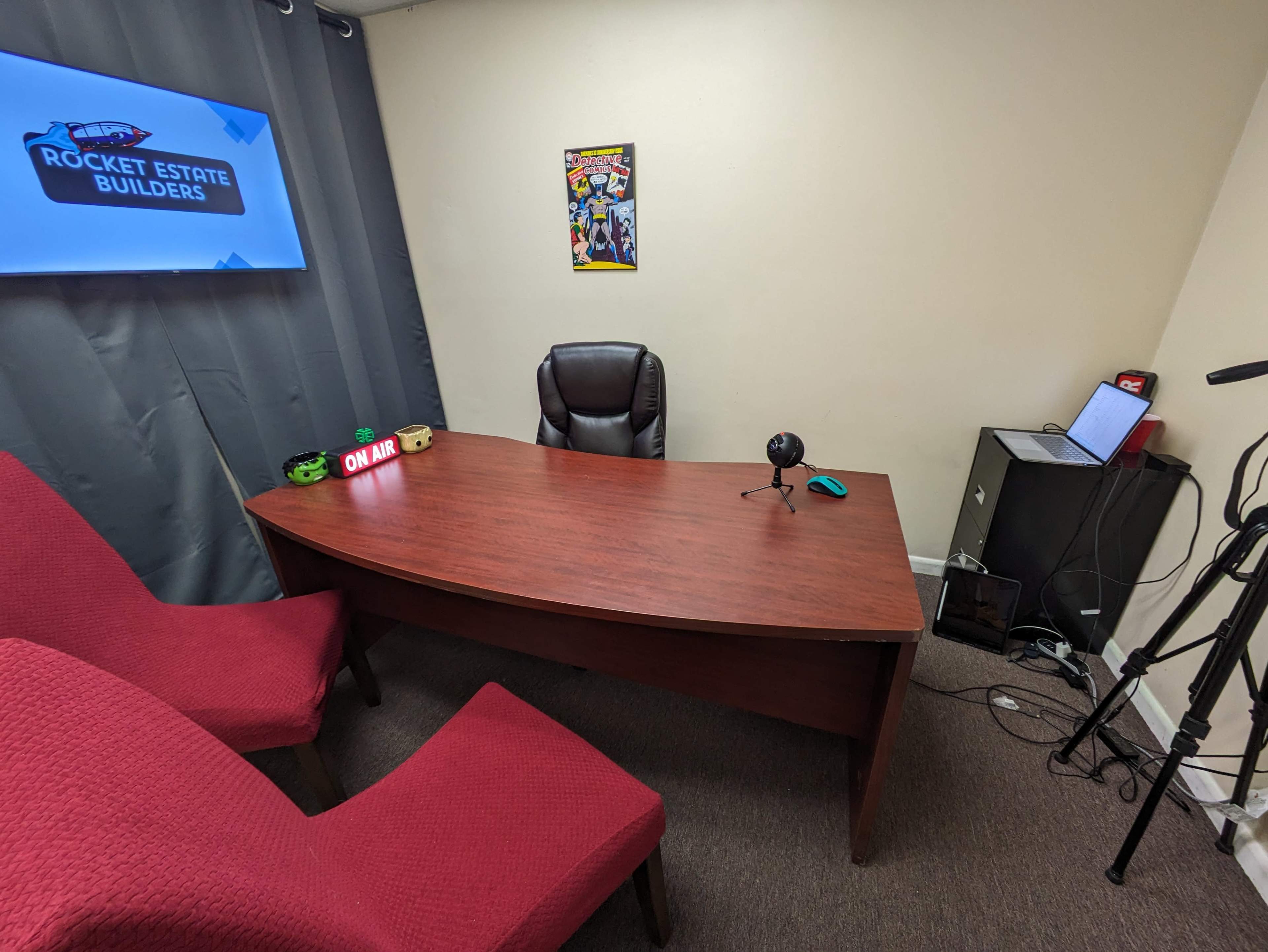 The image depicts a small office space featuring a wooden desk with a microphone, a laptop, and two red chairs, set against a gray curtain and a wall-mounted screen displaying a logo.