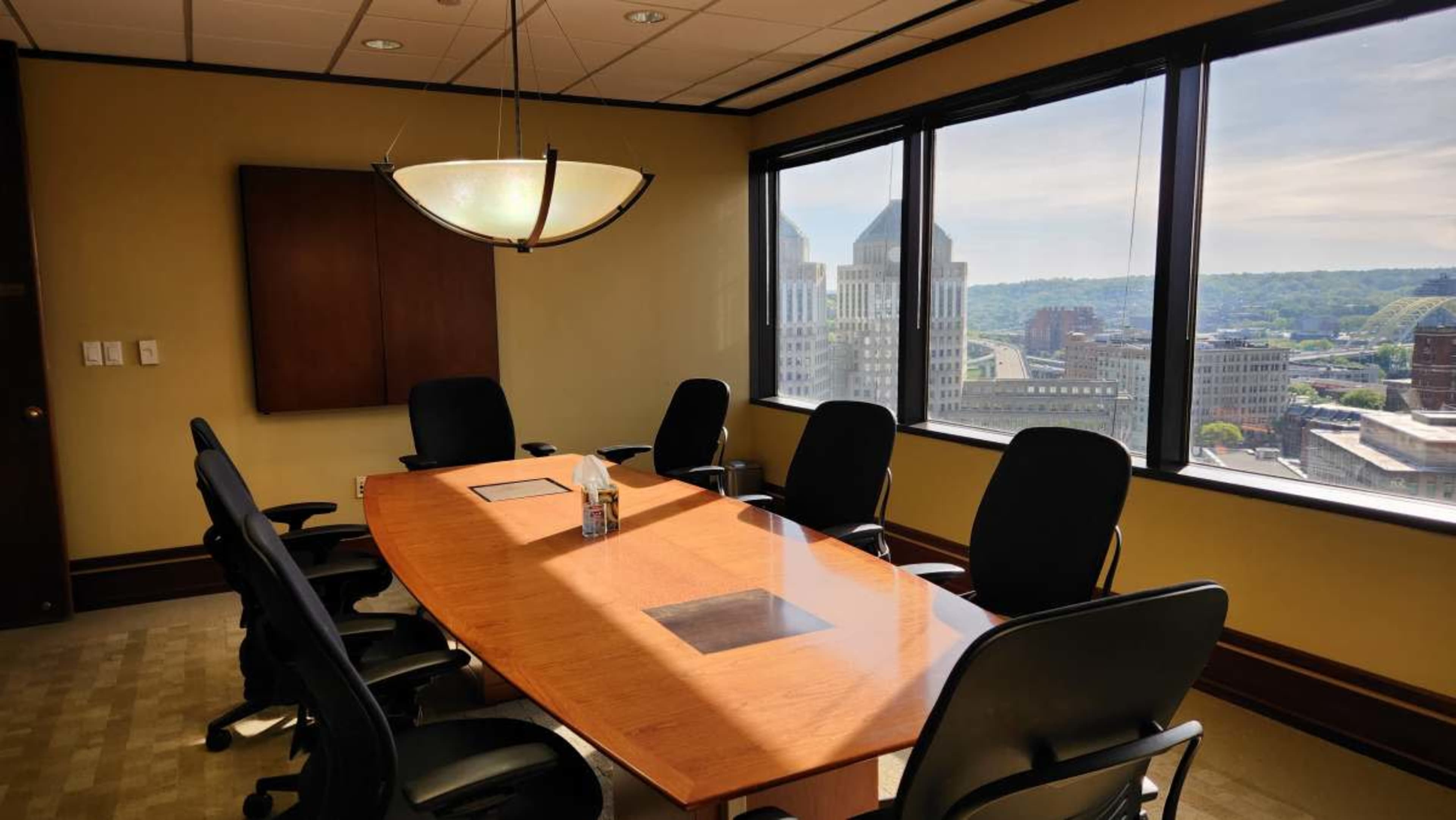 A conference room with a large wooden table surrounded by black chairs and a view of a cityscape through large windows.