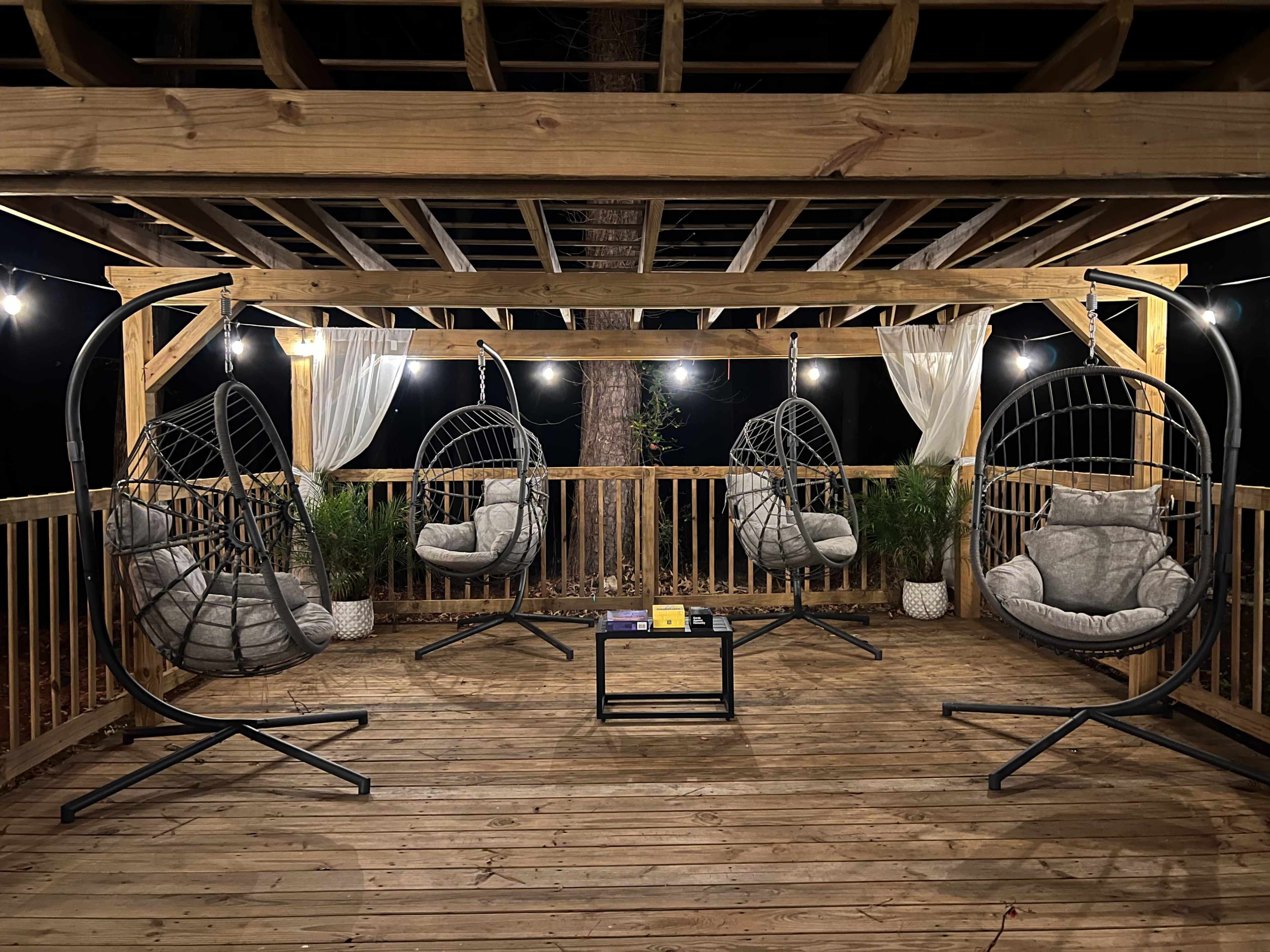 A wooden deck featuring four hanging chairs, a small table, and string lights illuminating the area at night.