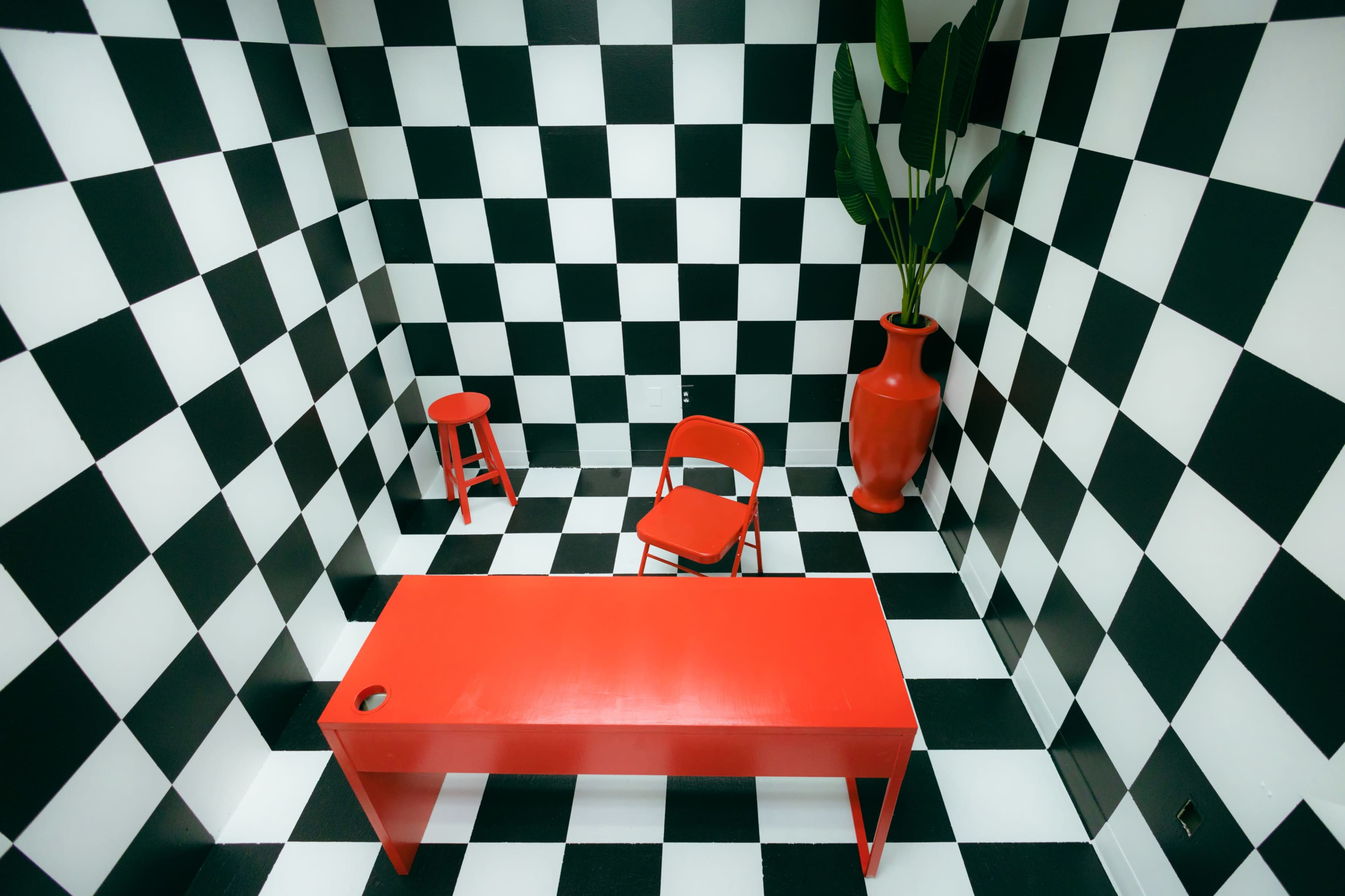 A room with black and white checkered walls and floor, featuring a red table, a red chair, a small red stool, and a large red vase with a green plant.