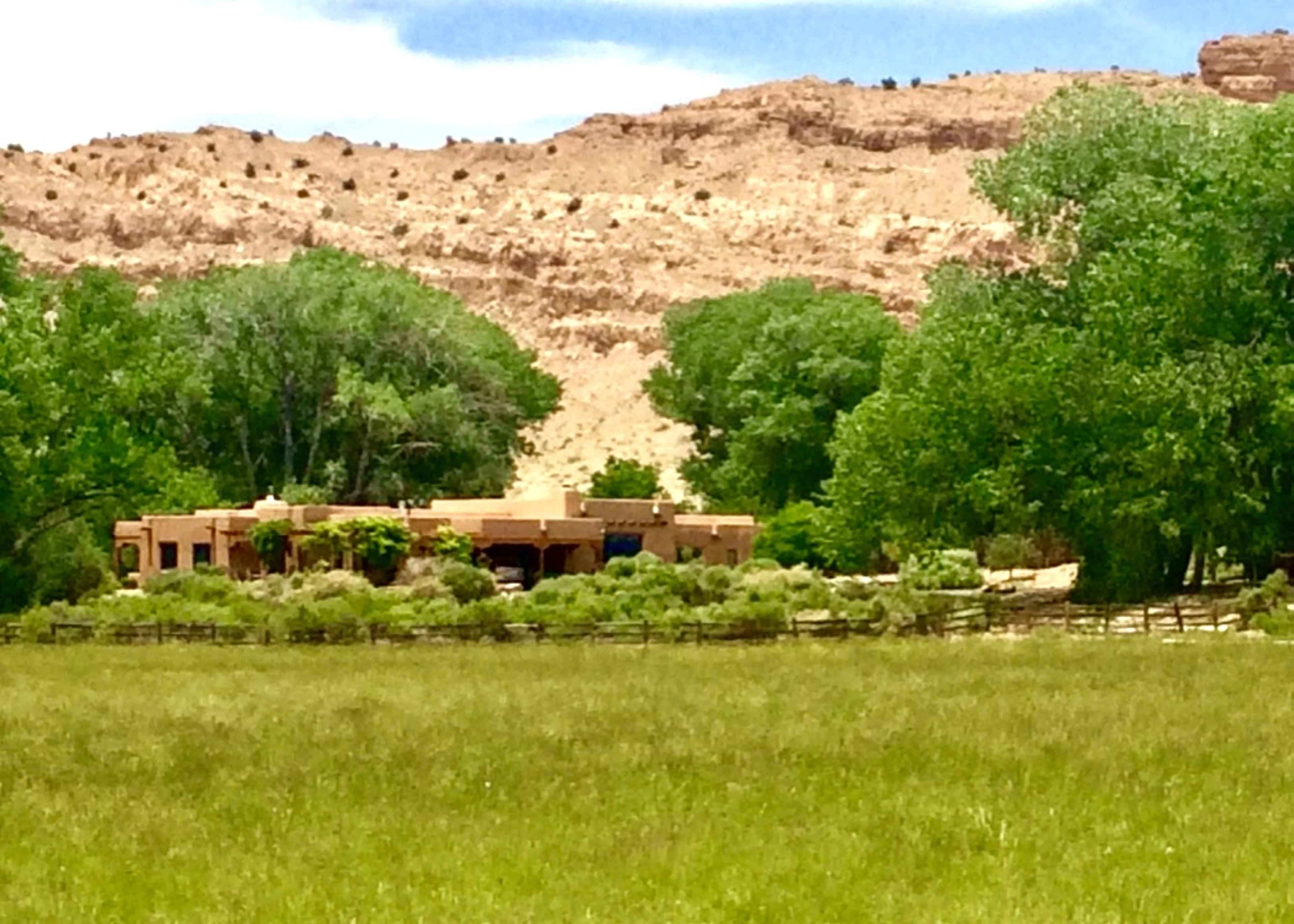A single-story adobe house is situated amid green trees and a grassy area, with rugged hills in the background.
