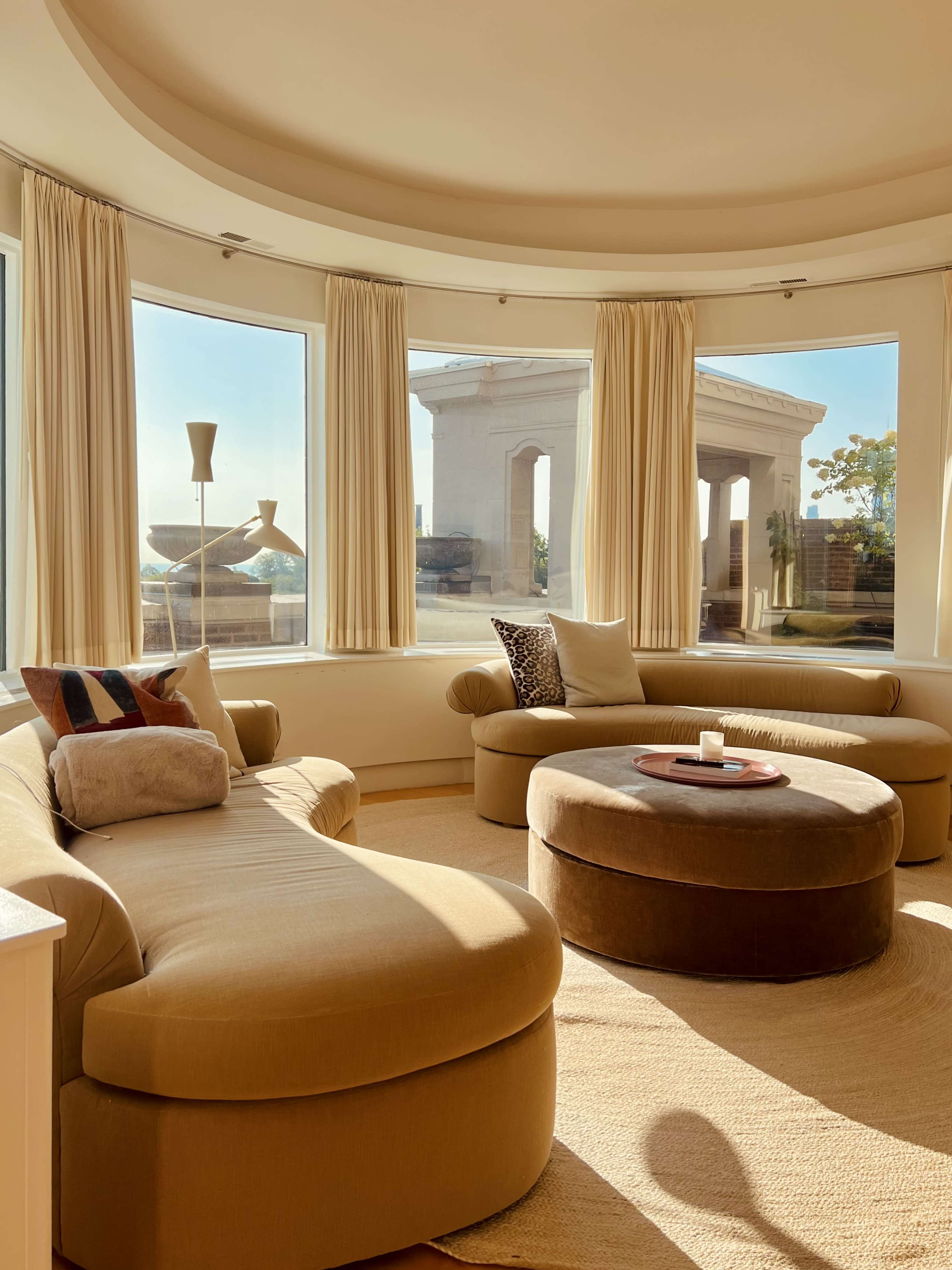 The image shows a cozy, modern living room with a curved sofa and a round coffee table, featuring large windows that let in natural light and provide a view of a structure outside.