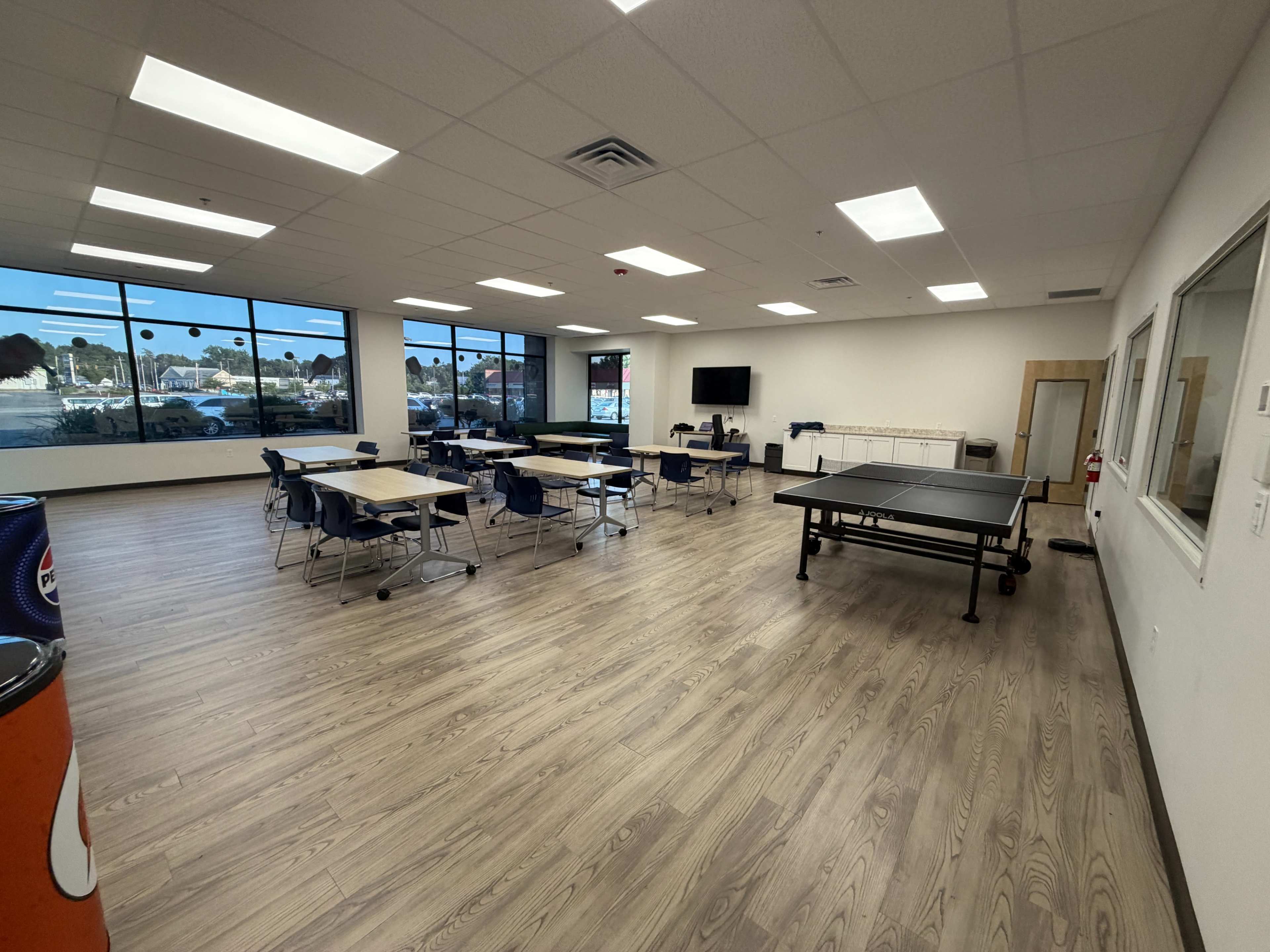 The image shows a spacious, well-lit room with tables, chairs, and a ping pong table, featuring large windows that overlook an outdoor area.