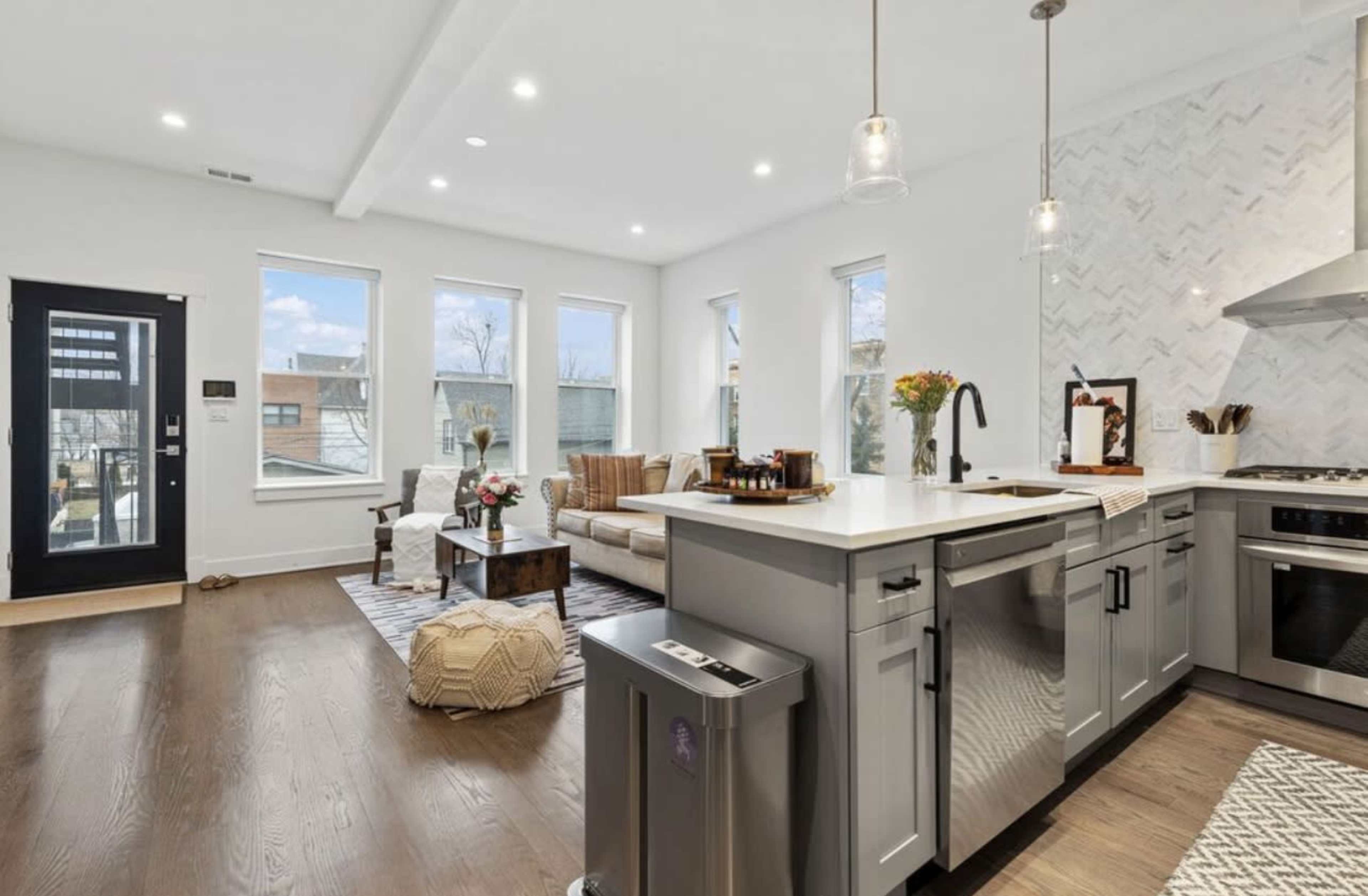 The image shows a modern open-concept kitchen and living room with large windows, light wood floors, and a sectional sofa.