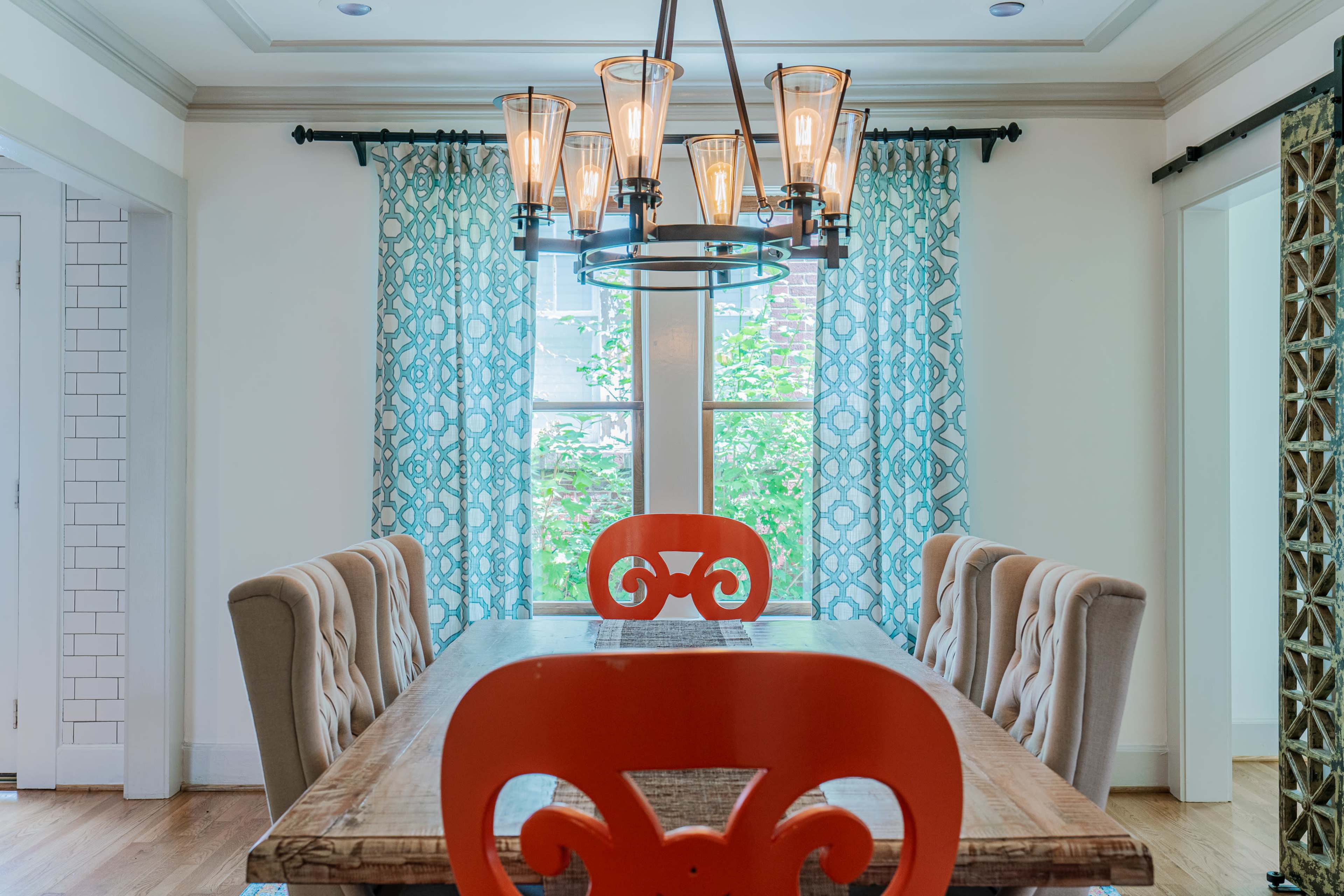 A dining room features a wooden table with an orange-accented centerpiece and upholstered chairs beneath a chandelier, flanked by blue patterned curtains over windows.