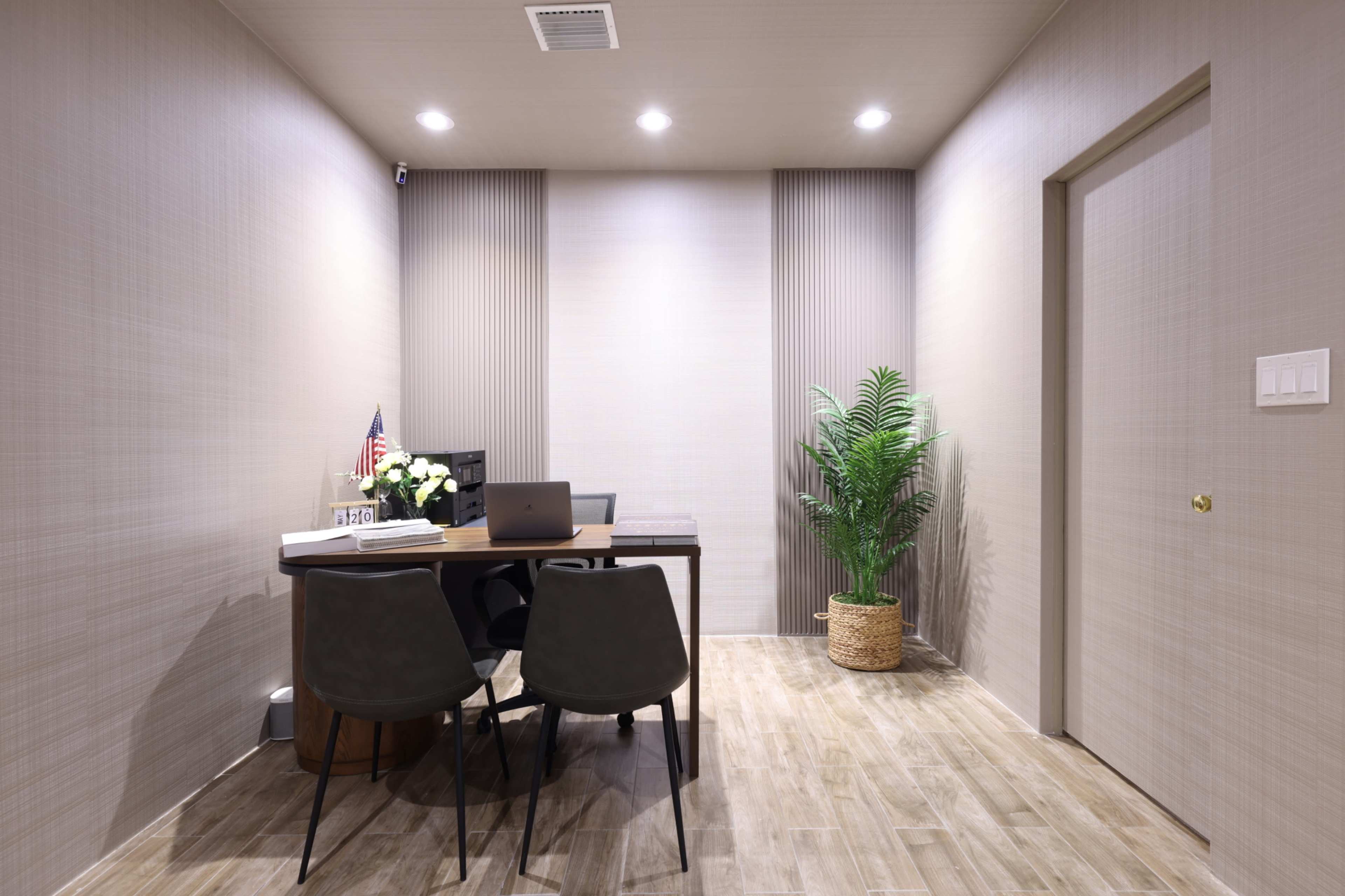 A small office space features a table with four chairs, a computer on the desk, and a potted plant in the corner.