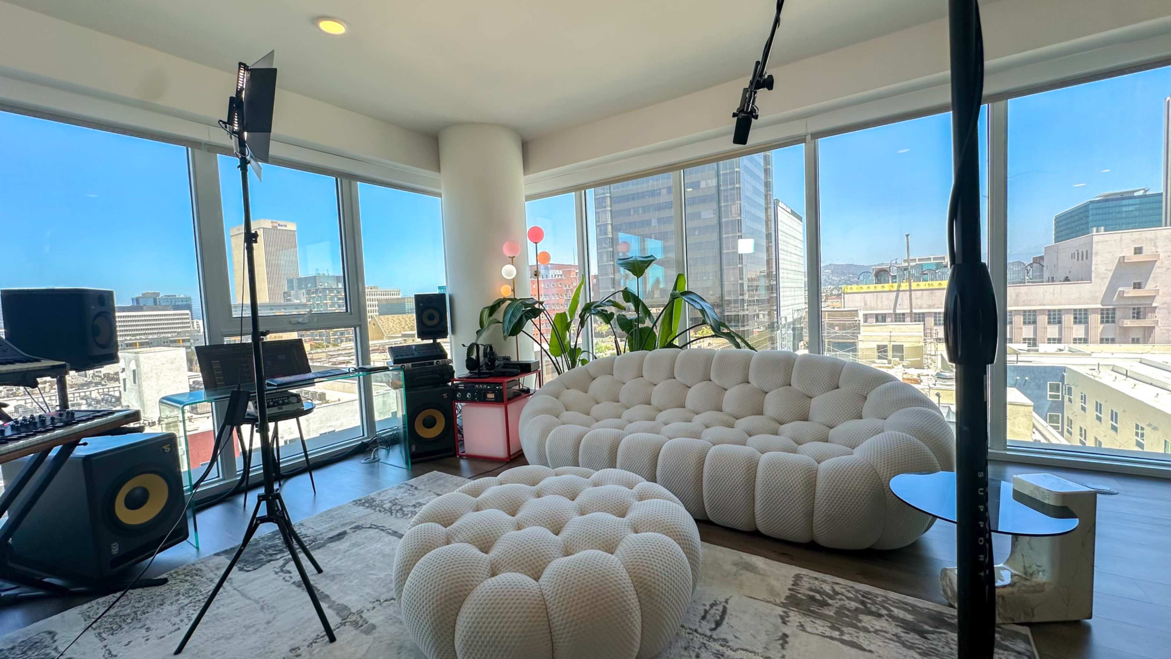 The image shows a modern studio space with large windows revealing a cityscape, featuring a plush white couch, a round table, and audio equipment.