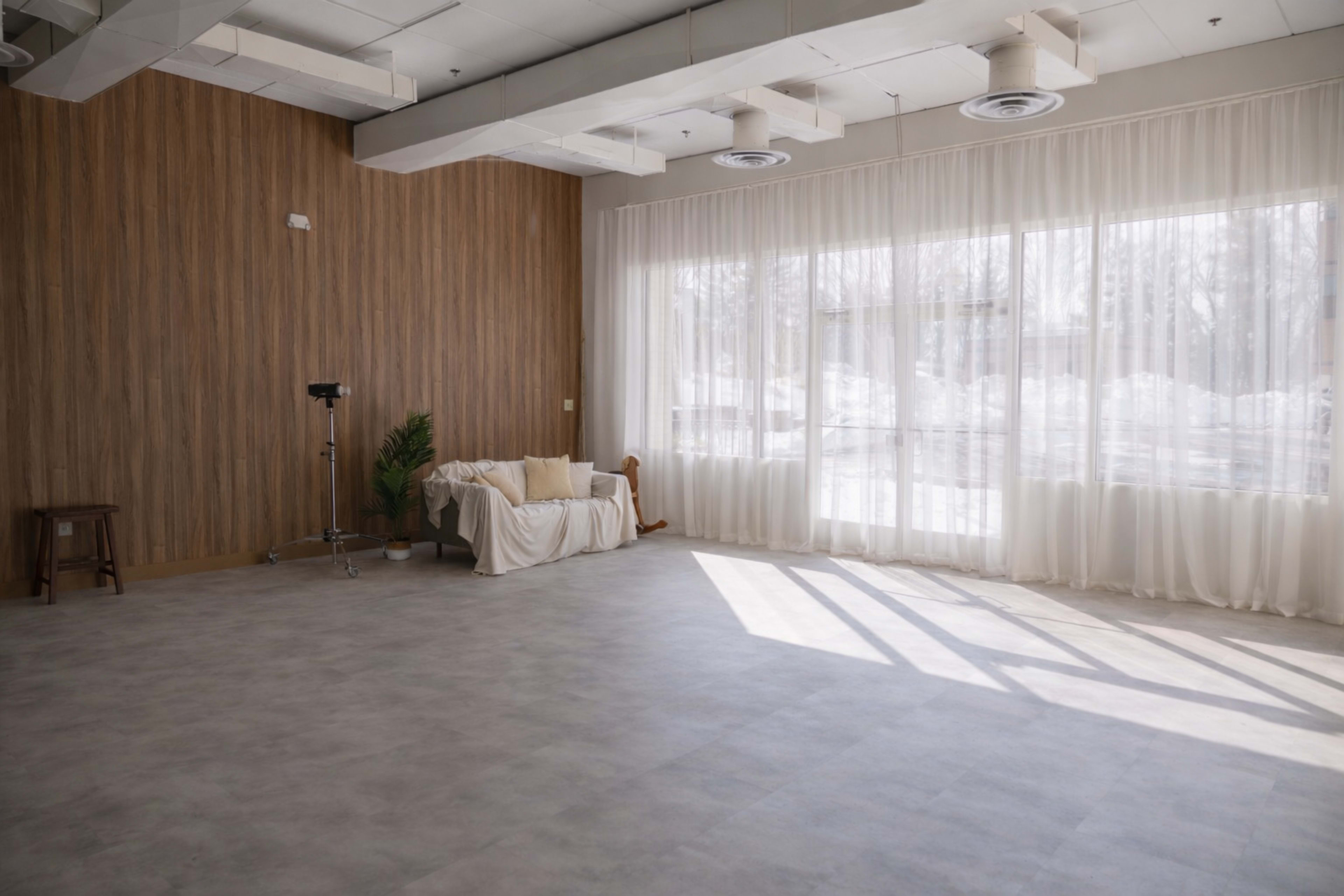 The image shows a spacious, brightly lit room with a large window, light-colored flooring, and a white couch adorned with pillows.