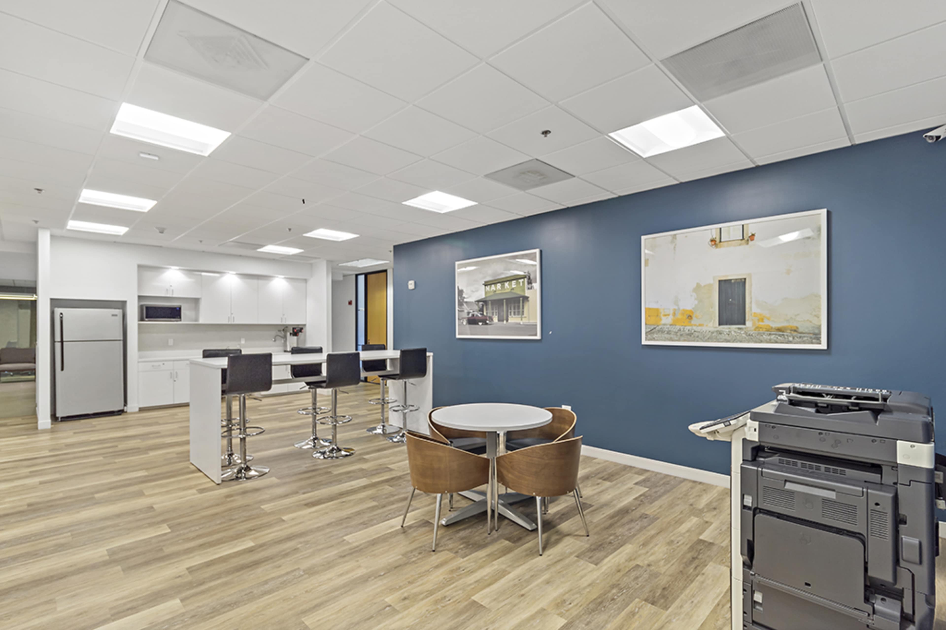 A modern office break area featuring a kitchenette with bar stools, a round table with chairs, and a printer station against a blue accent wall.