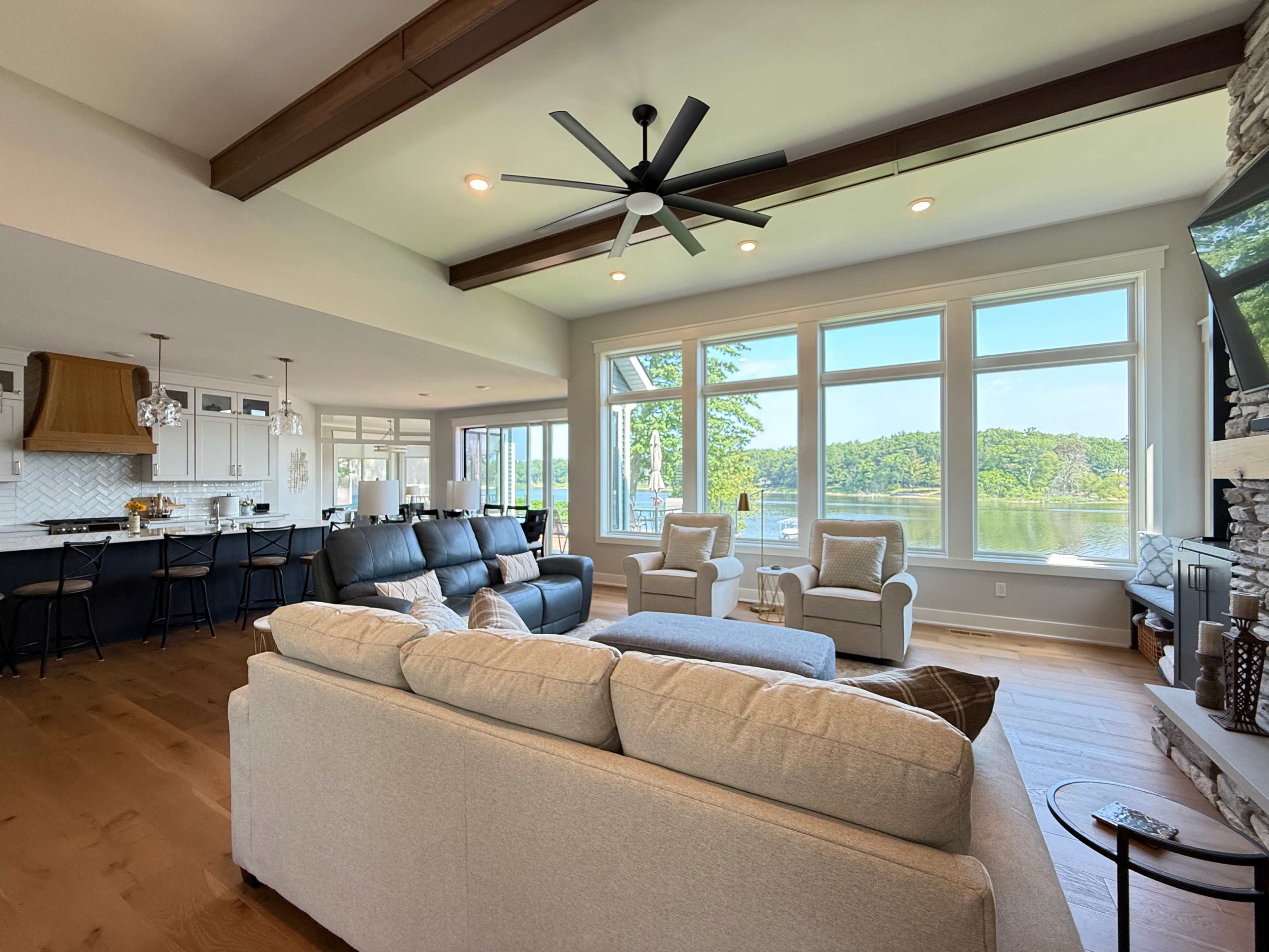 The image shows a modern living room with large windows overlooking a lake, featuring a comfortable seating area and a kitchen in the background.