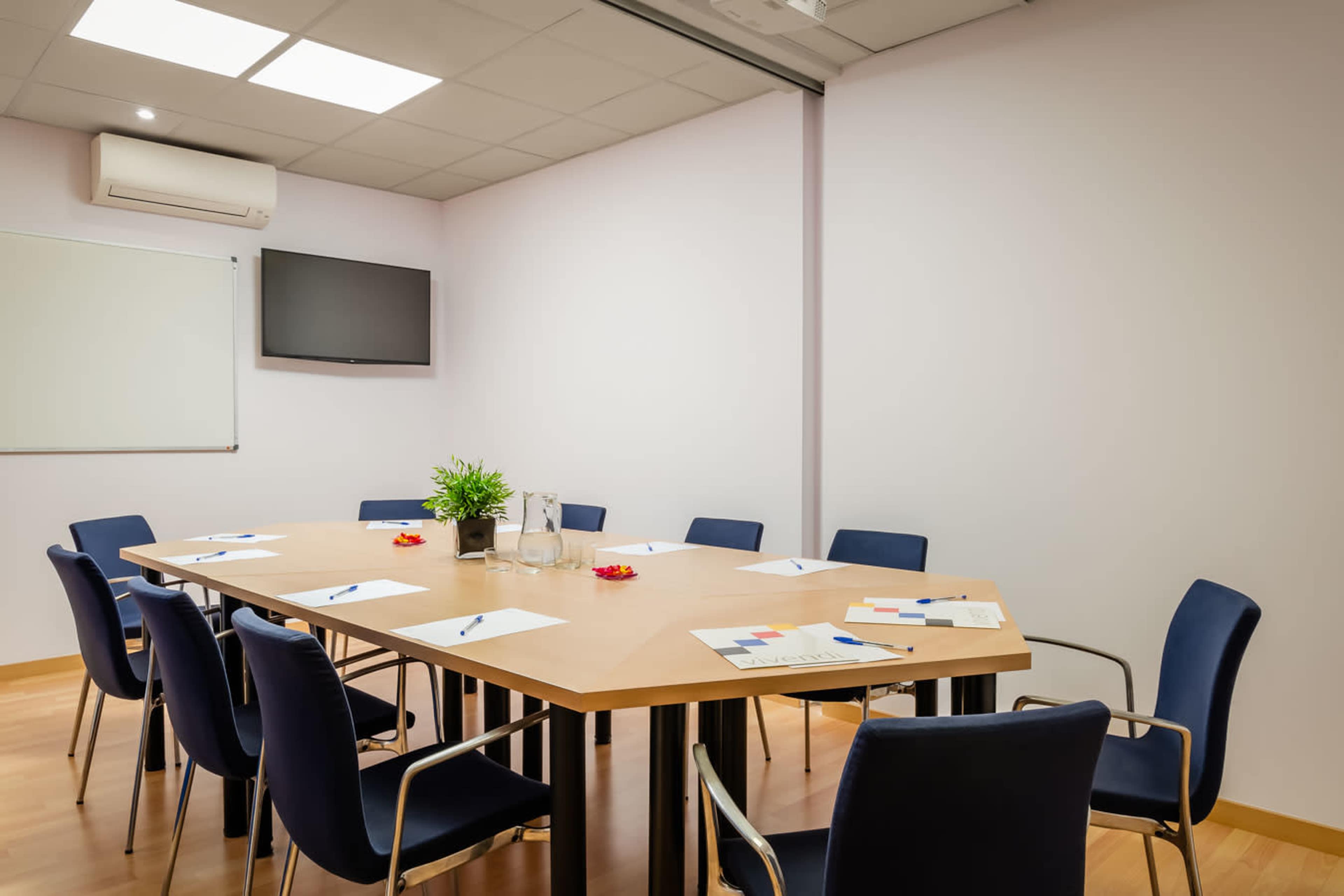 A conference room features a large table surrounded by eight chairs, with a television mounted on the wall and a whiteboard in the corner.