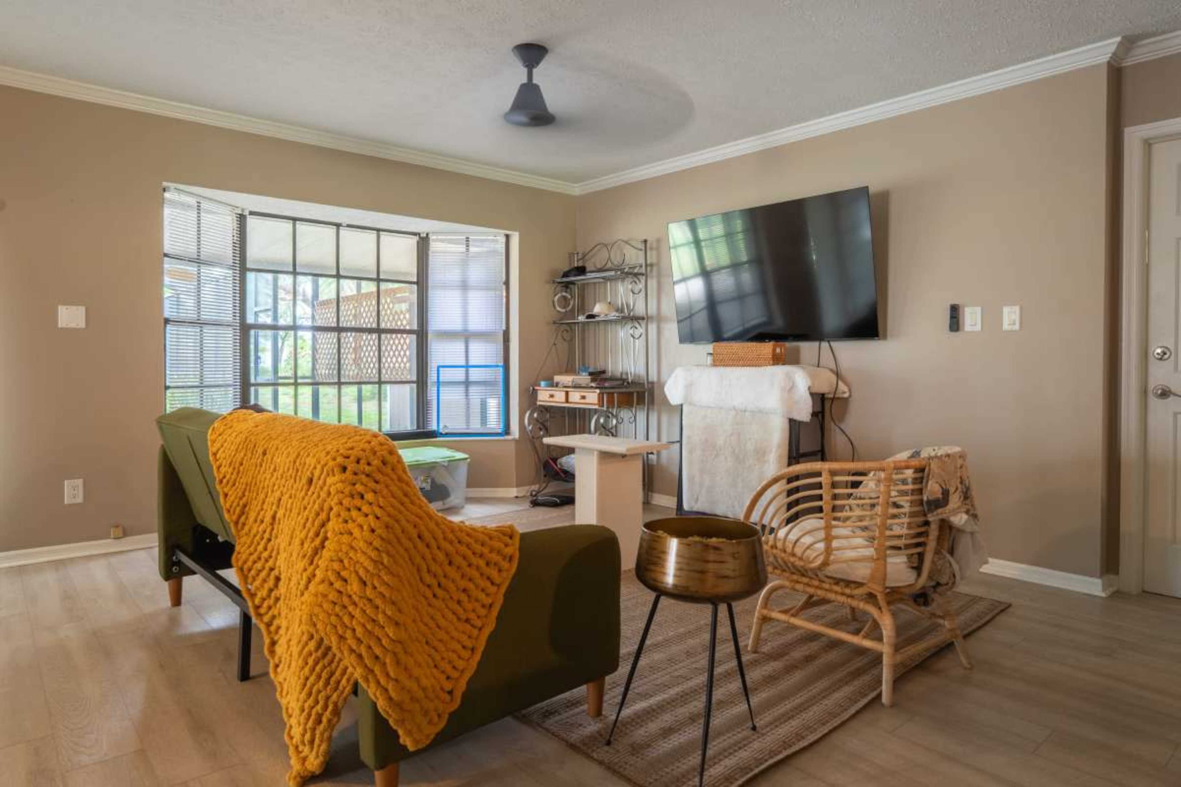 The image shows a cozy living room featuring a large window, a television mounted on the wall, a green sofa with a yellow throw blanket, a wooden coffee table, and a rattan chair.