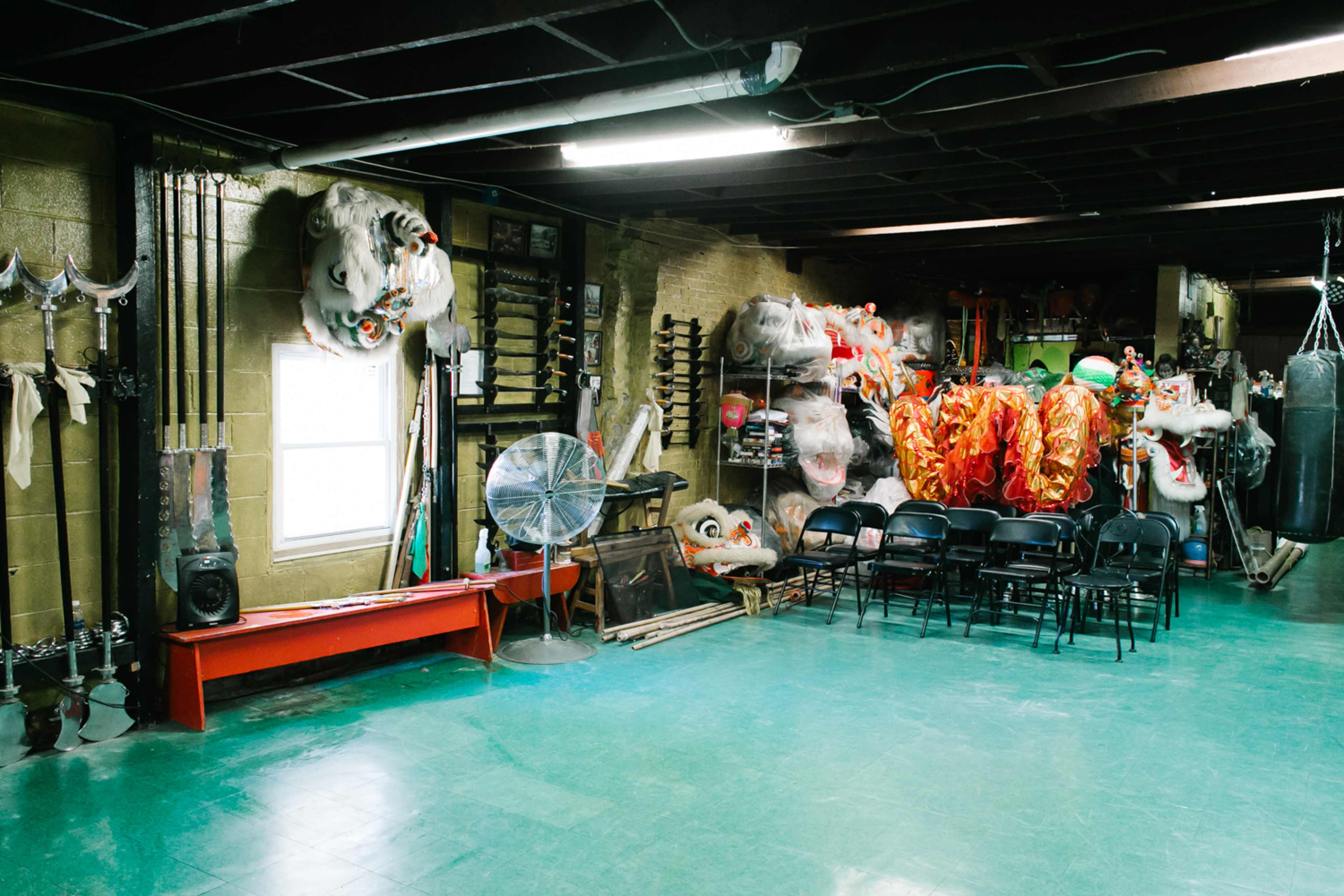 An indoor space with martial arts weapons on the walls, a fan in the corner, and colorful dragon and lion costumes hanging next to empty chairs.