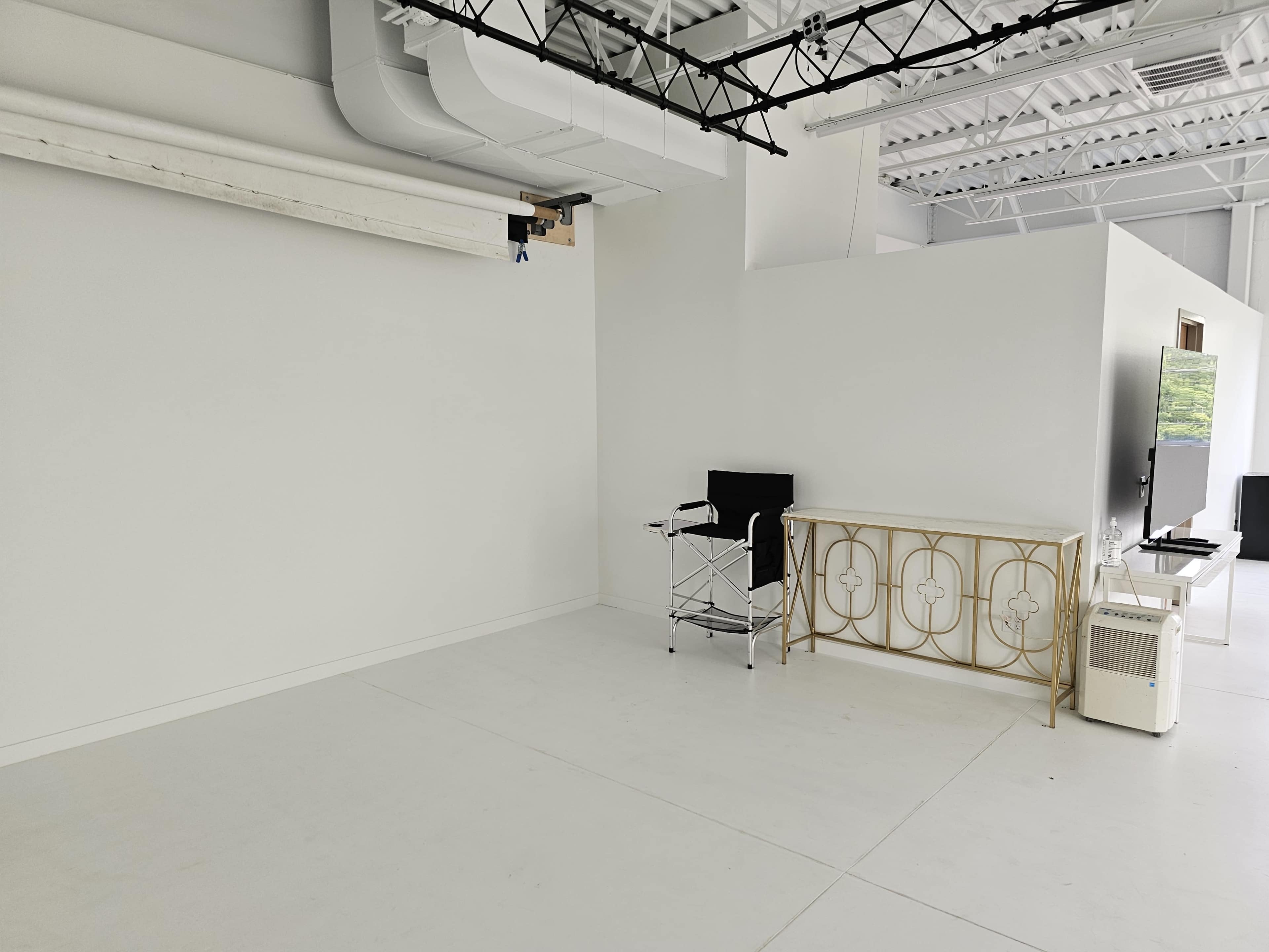 The image shows an empty room with a white floor, white walls, a small table with a decorative design, a black chair, and a television mounted on the wall.