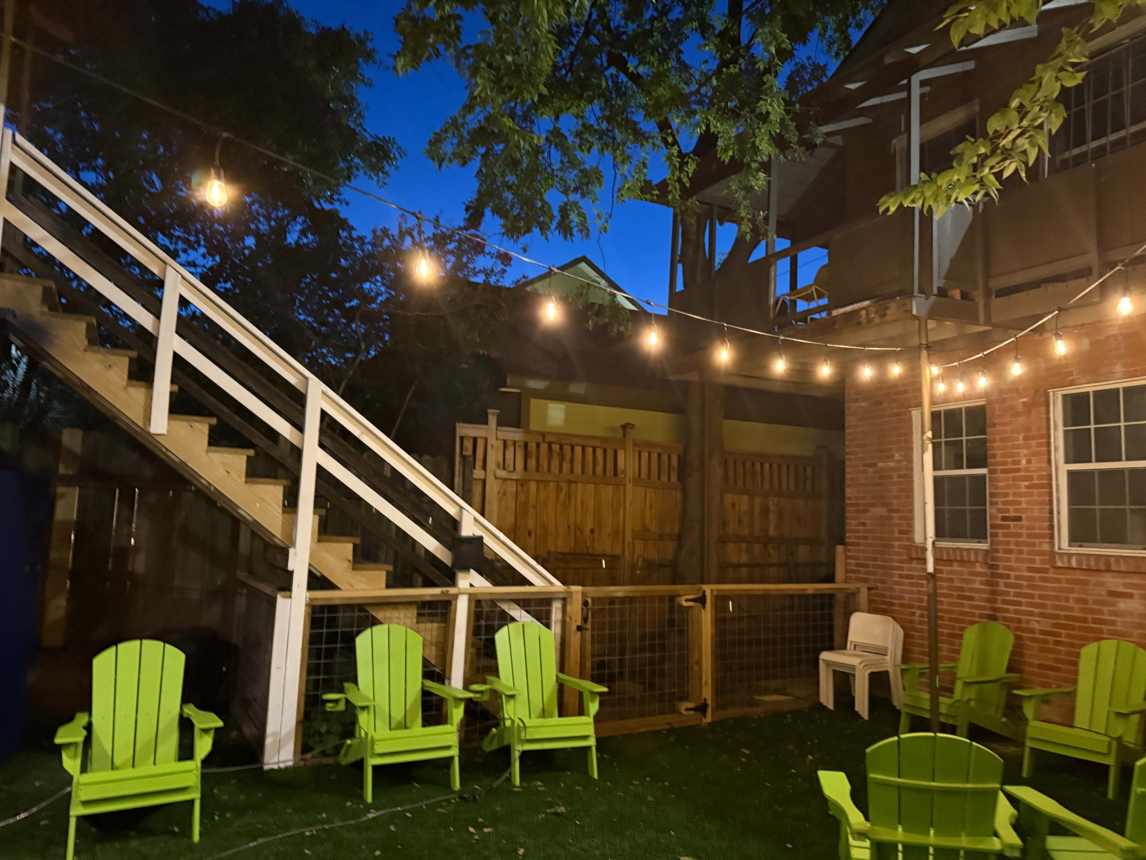 The image shows a backyard scene at dusk with green Adirondack chairs, string lights overhead, and a wooden staircase leading to a building in the background.