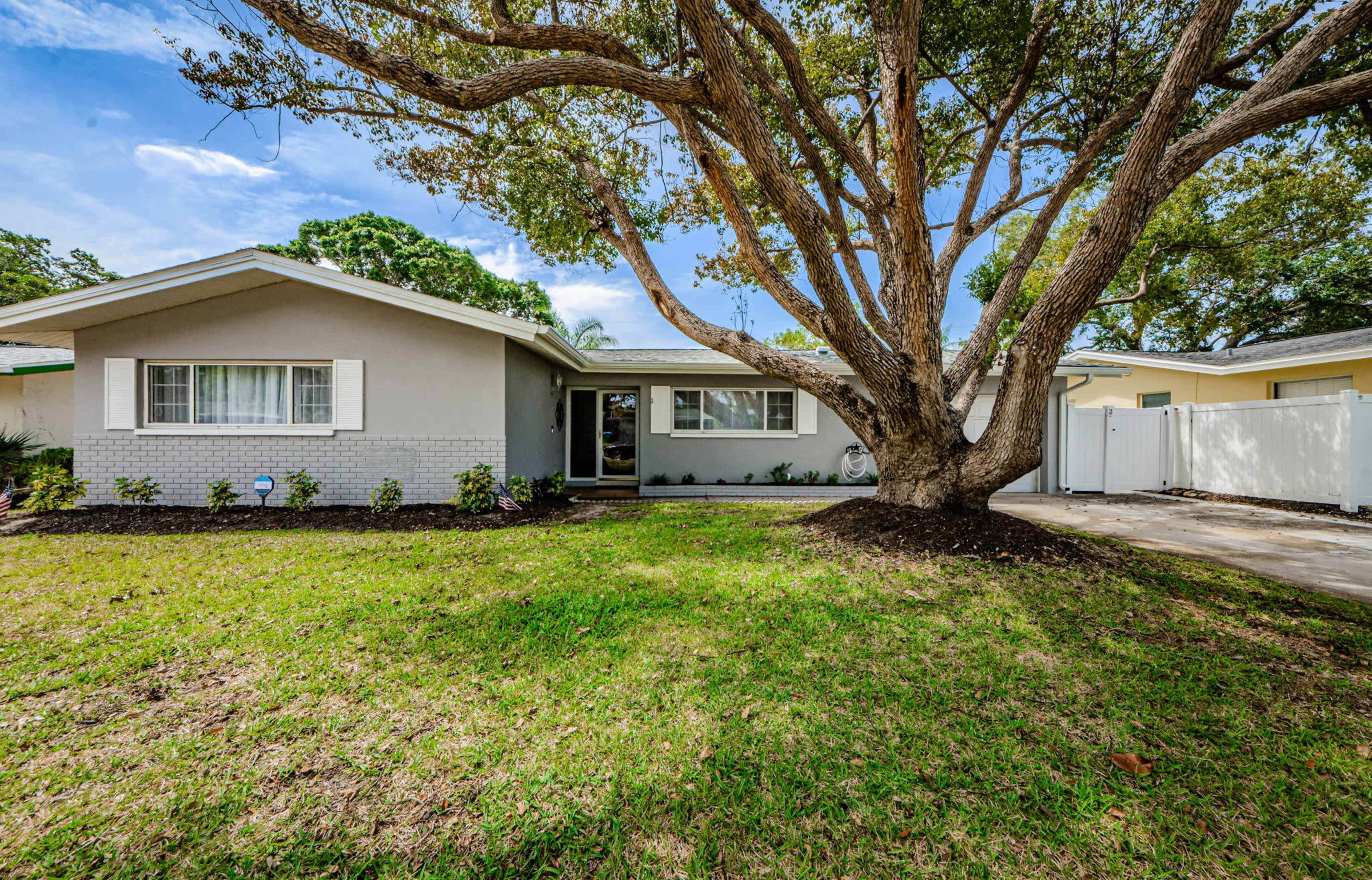 A single-story house with a large tree in the front yard is surrounded by grass and landscaping.