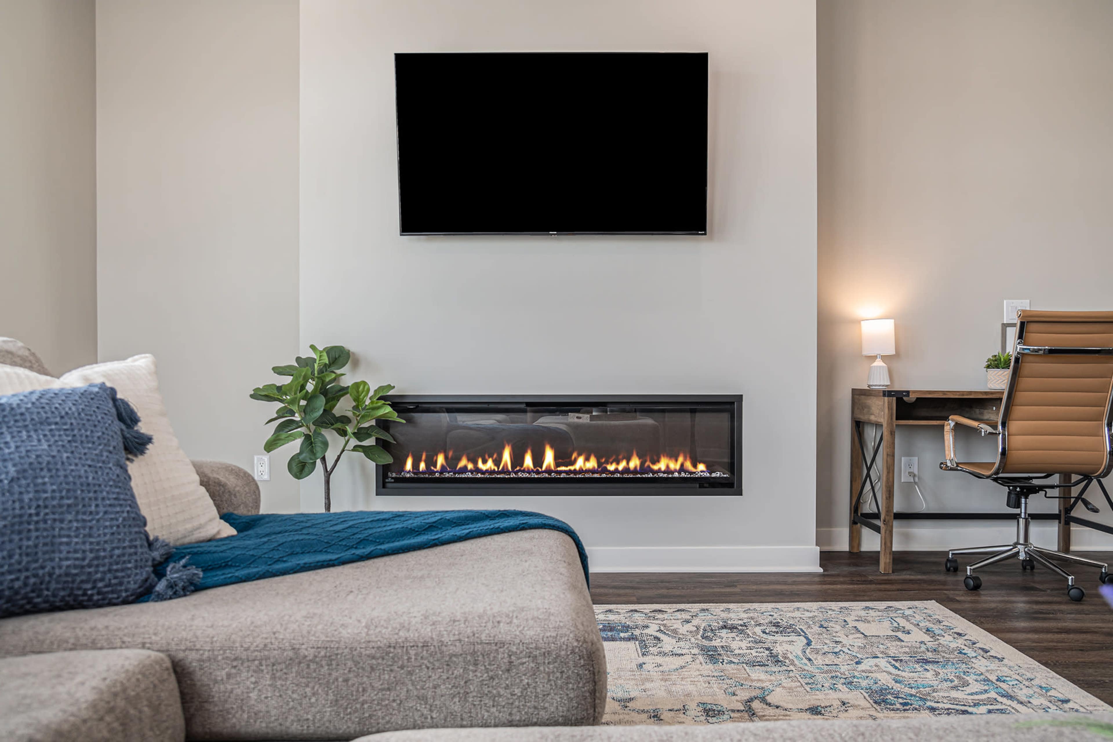 A modern living room features a wall-mounted television above a sleek fireplace with a glass front, accompanied by a desk and a chair in the corner.