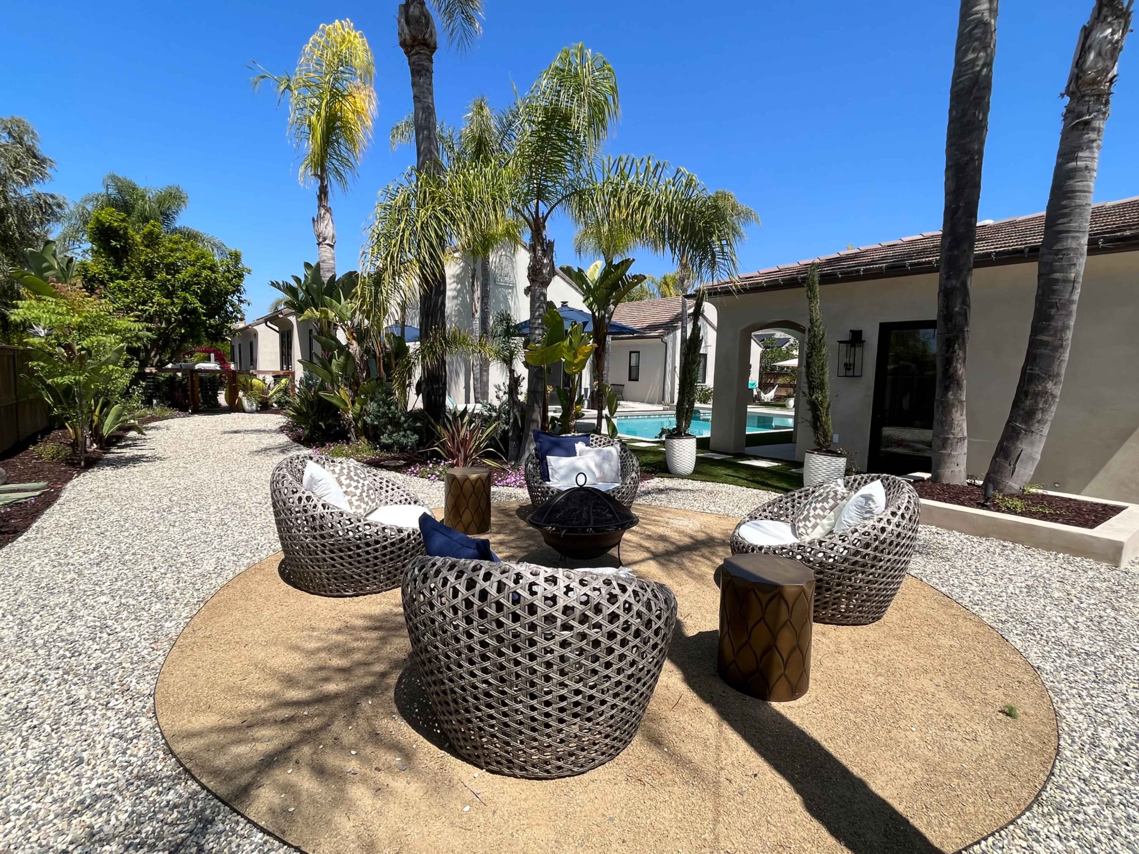 A circular seating area with four woven chairs and a central fire pit is surrounded by palm trees and landscaped greenery.