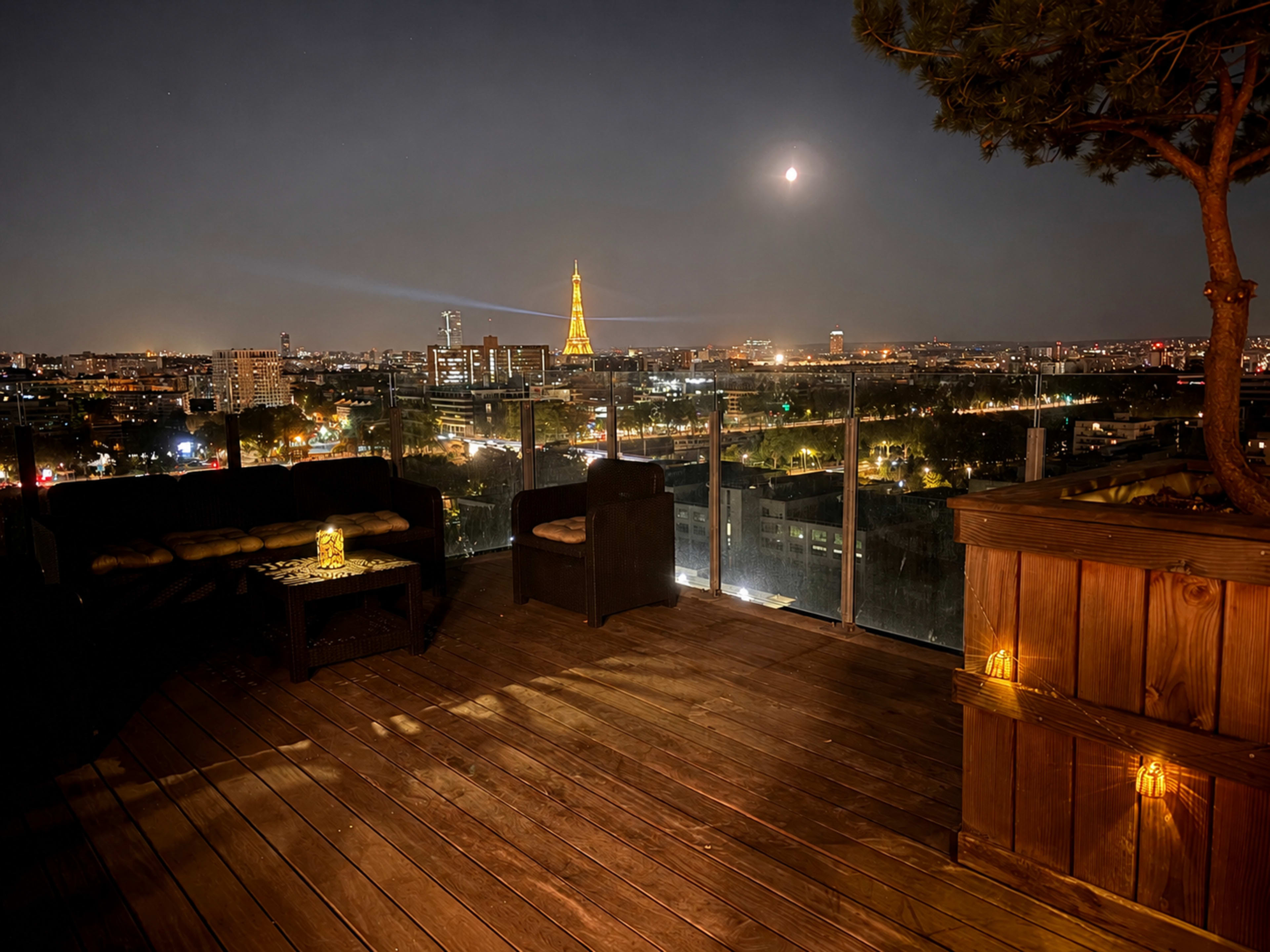 A rooftop terrace with wooden flooring offers a view of the illuminated Eiffel Tower and a city skyline under the night sky, featuring a full moon.
