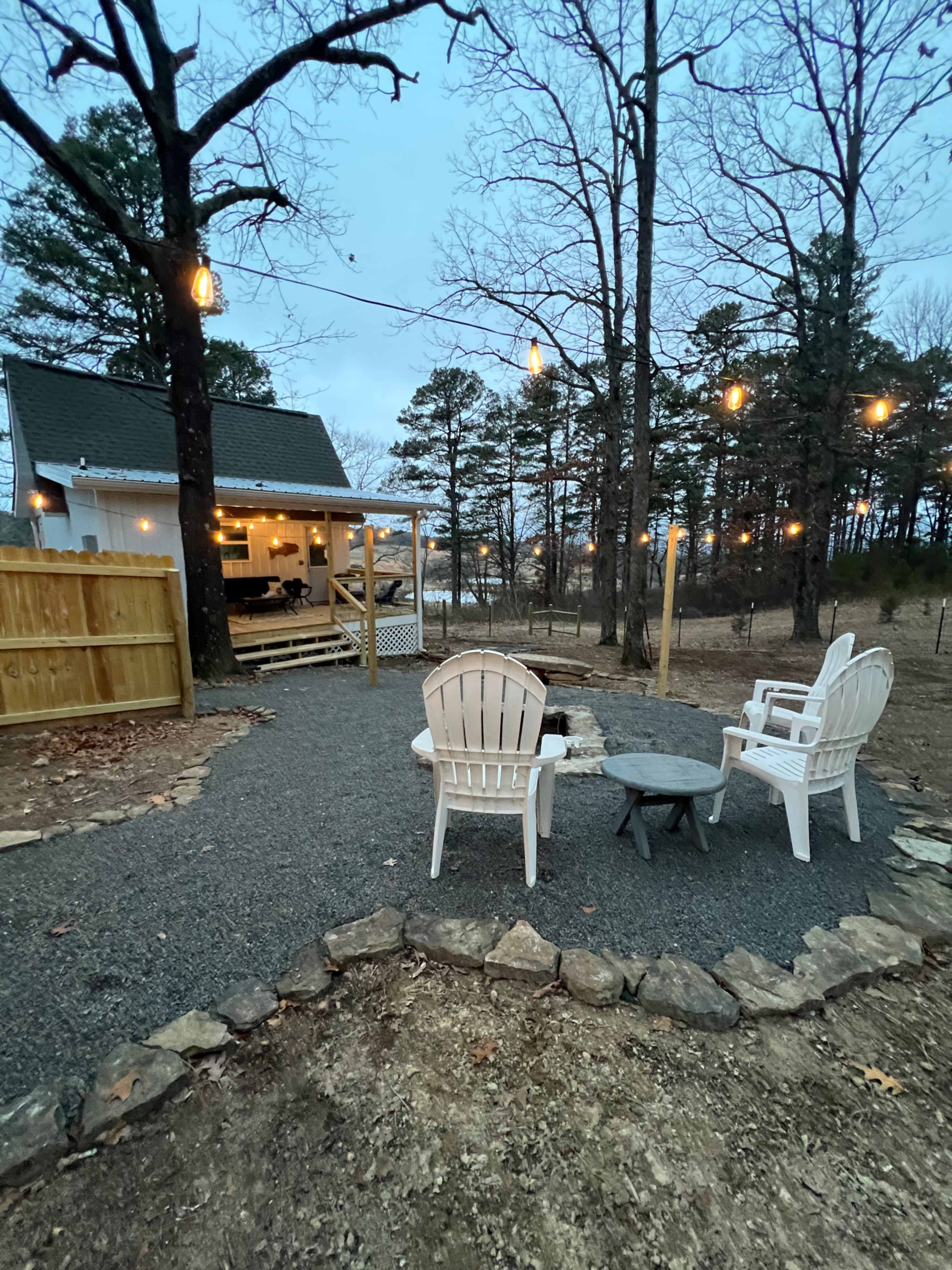 A cozy outdoor seating area with white chairs and a fire pit, surrounded by trees and leading to a house with string lights.