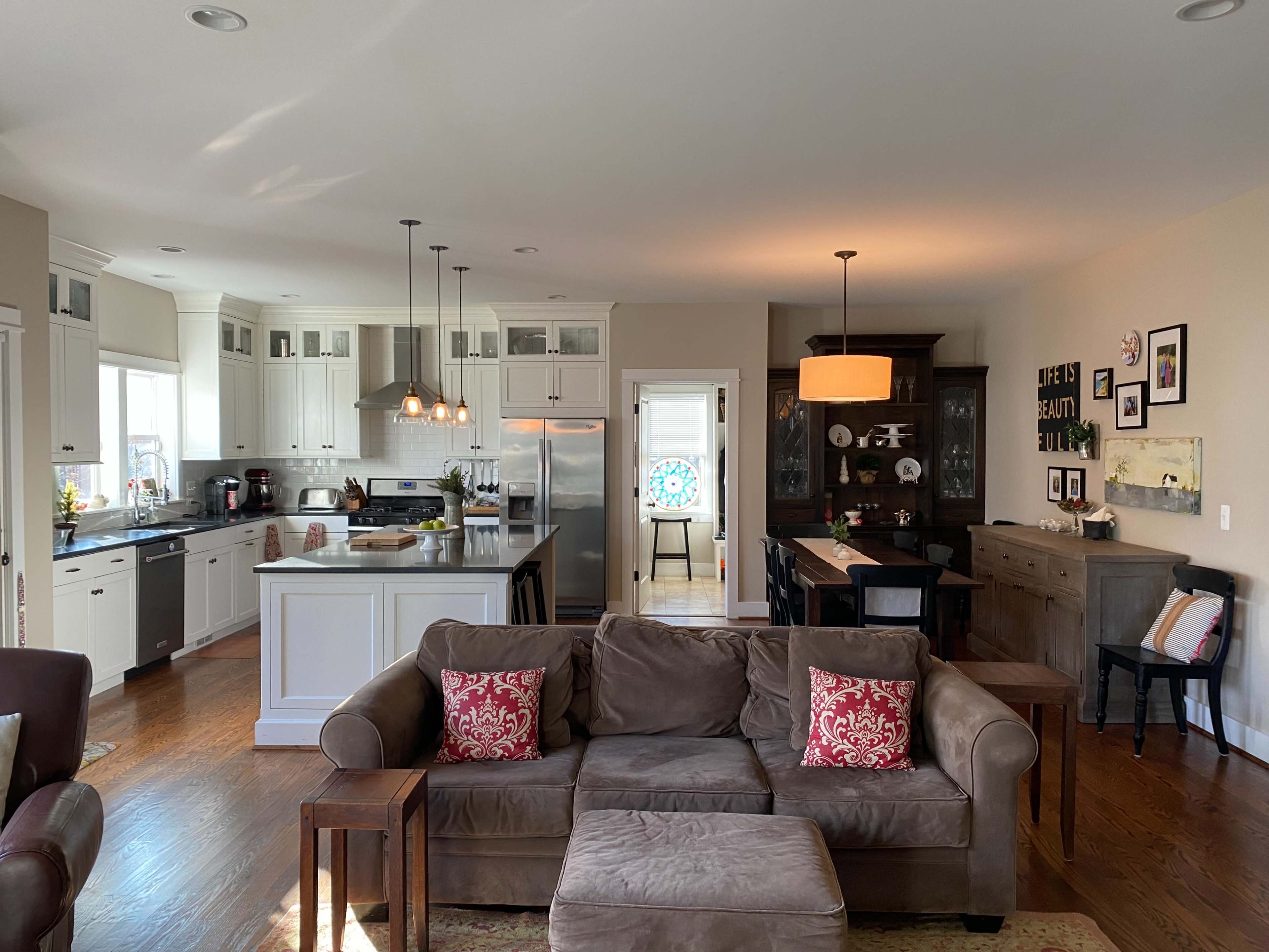 The image shows a bright, open-plan living area with a sofa, dining space, and kitchen featuring white cabinetry and a wooden table.