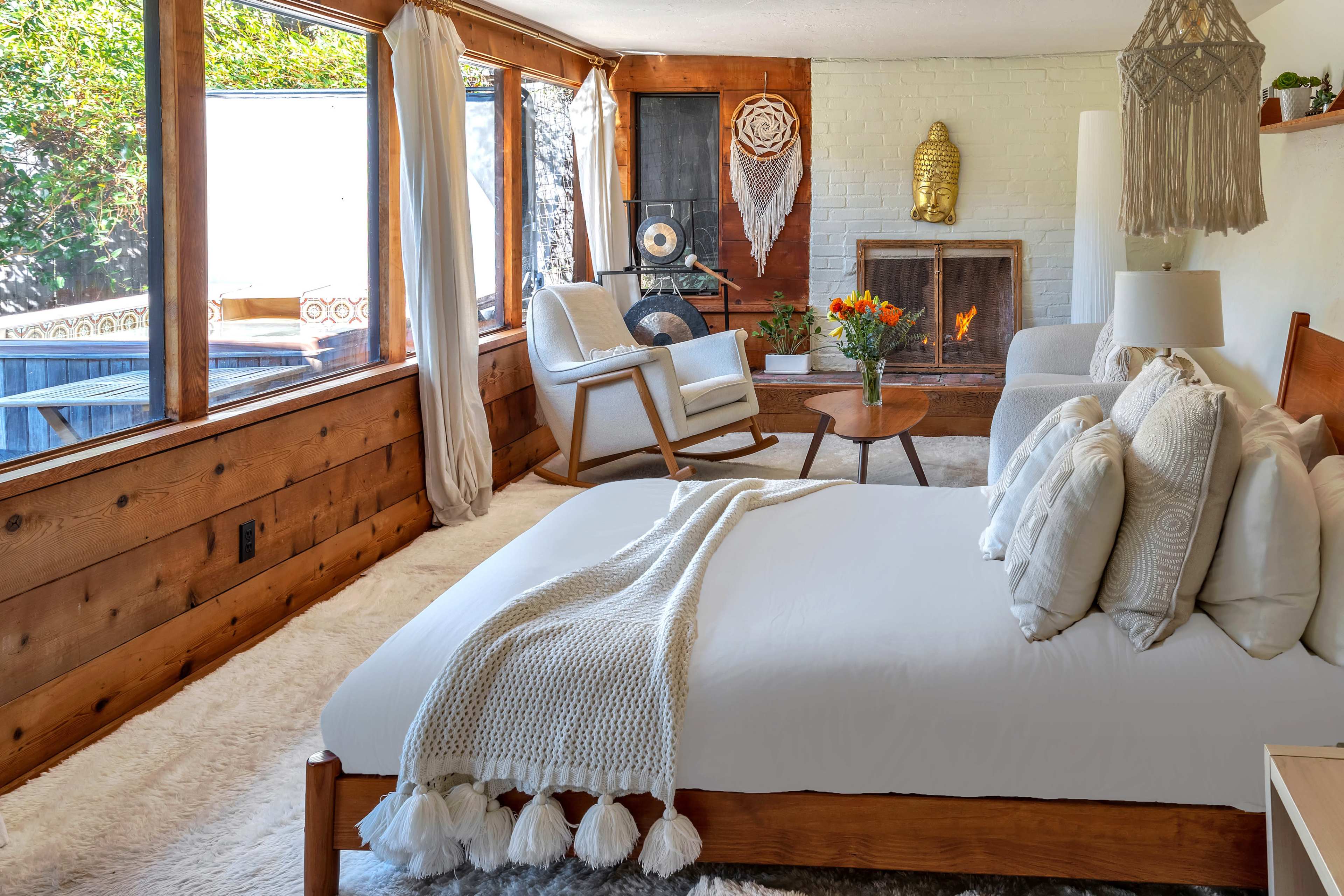 The image shows a cozy, light-filled room featuring a bed, a fireplace, and seating areas with wooden accents and decorative items.