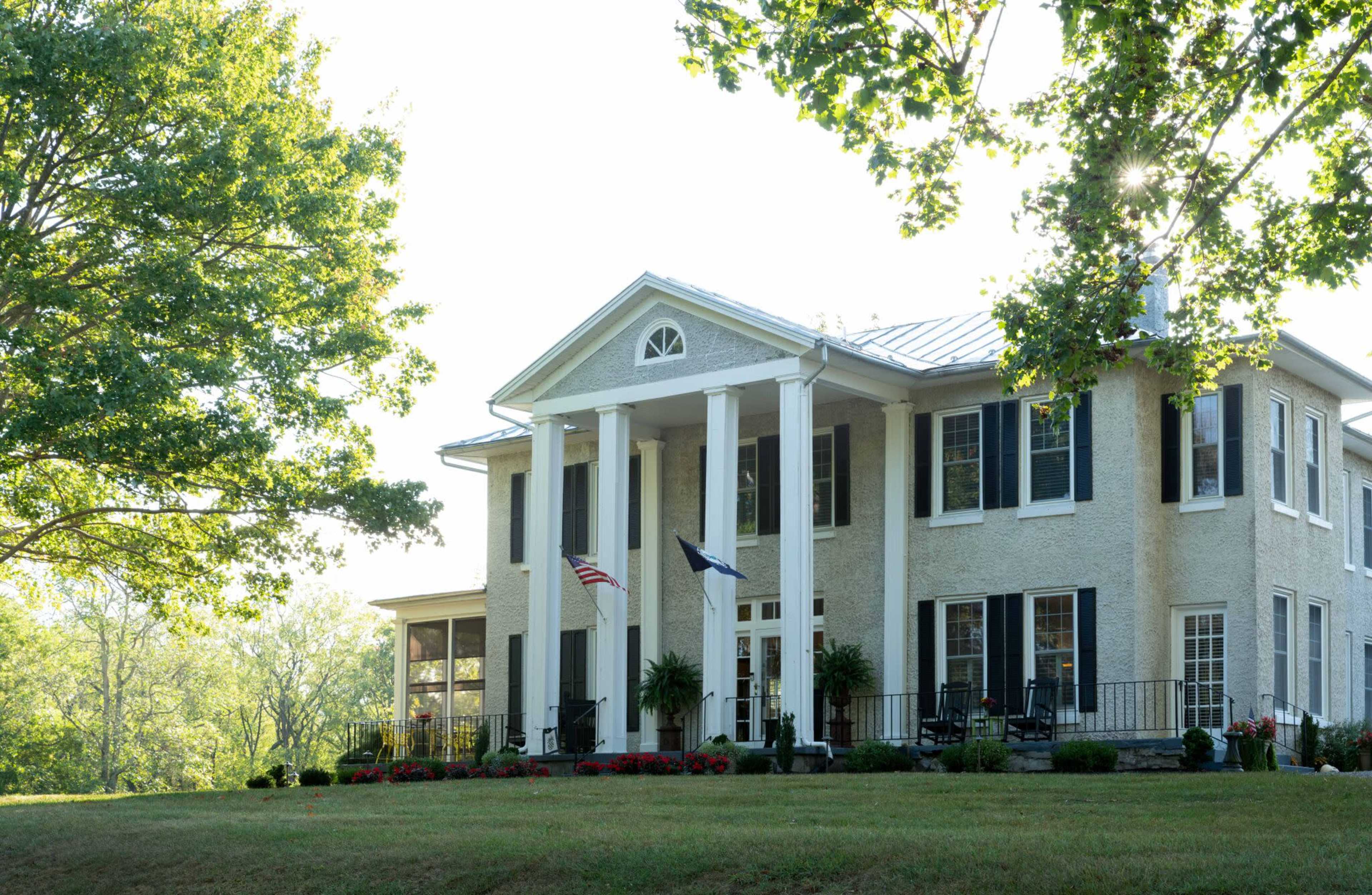 A large, two-story house with white columns, surrounded by greenery and flower beds, and two flags displayed in front.