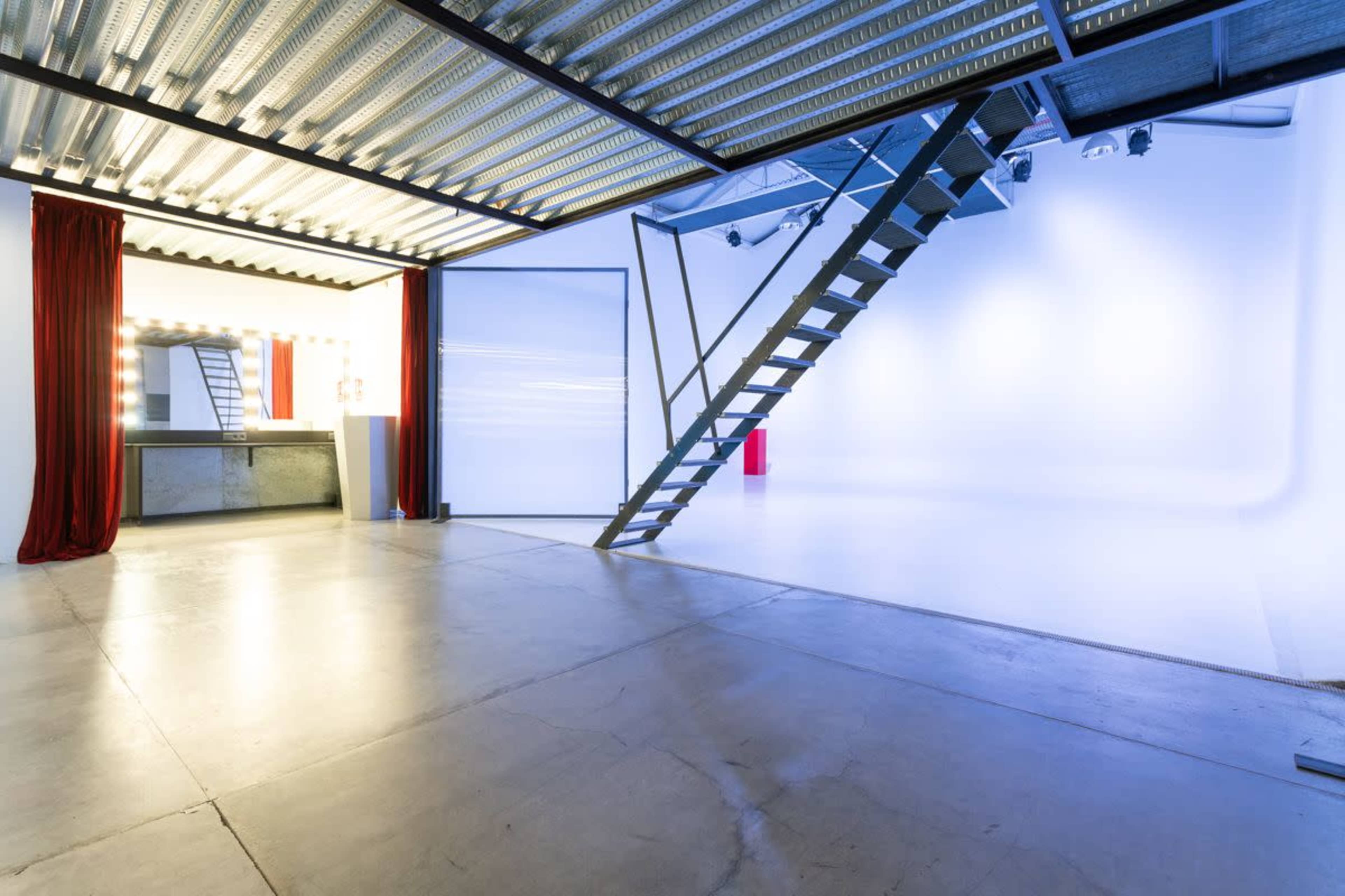 The image shows a spacious, modern studio with a staircase leading to a second level, featuring a vanity area with lights and a stark, well-lit white backdrop.