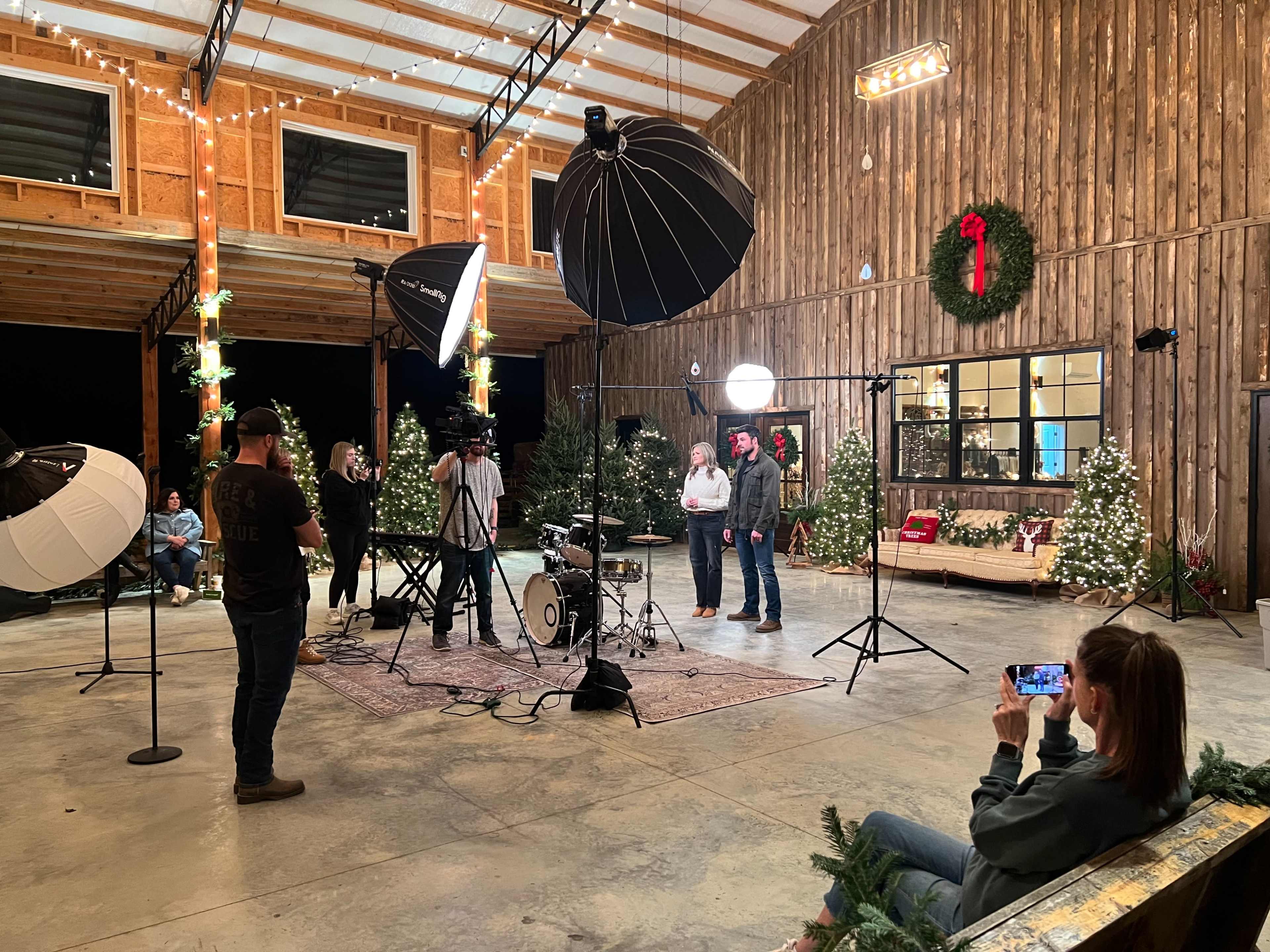 A group of people gathers around a photography setup inside a decorated barn with Christmas decorations and studio lighting equipment.