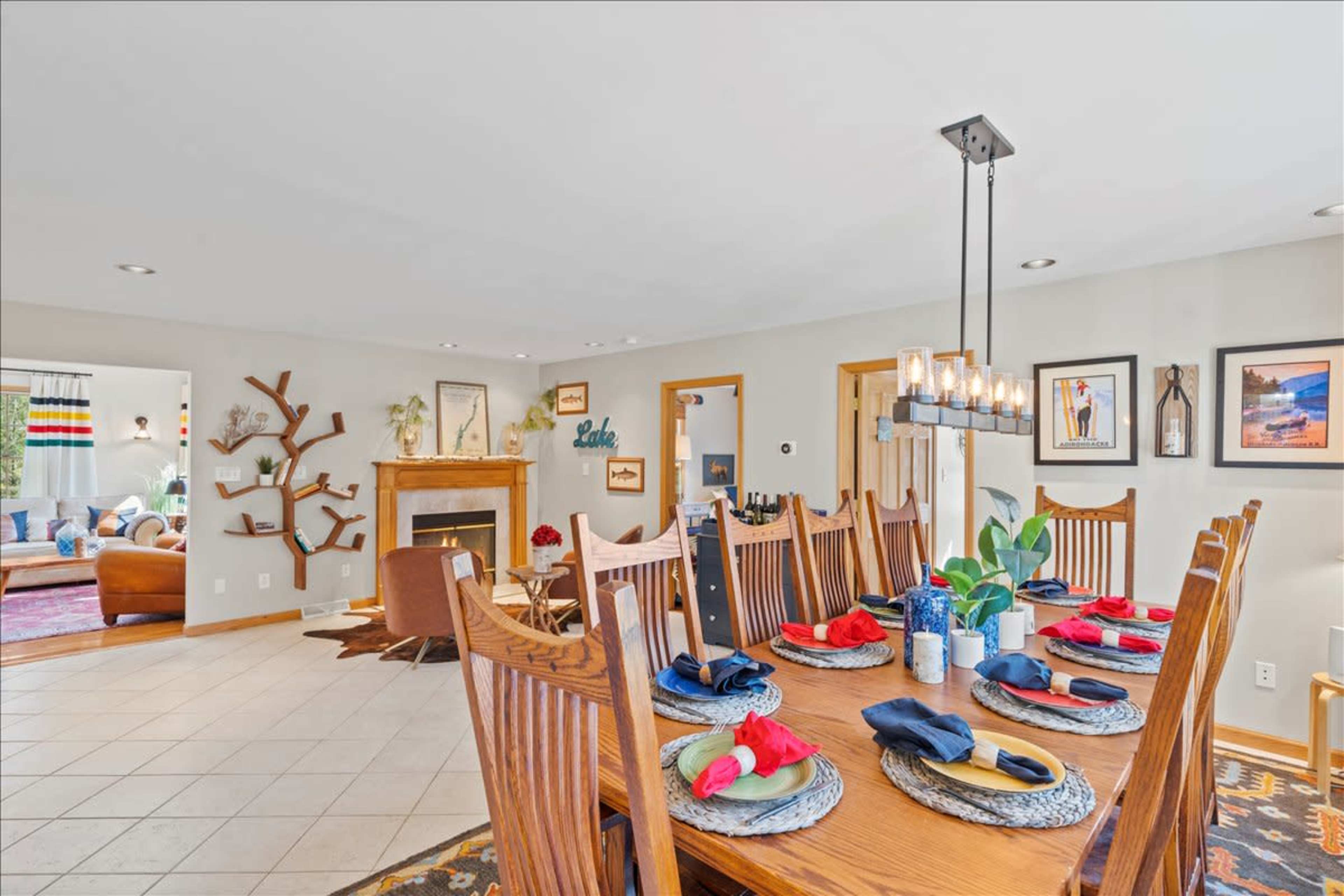 A dining room with a large wooden table set for a meal, featuring placemats, plates, and decorative napkins, adjacent to a living area with a fireplace and artwork on the walls.