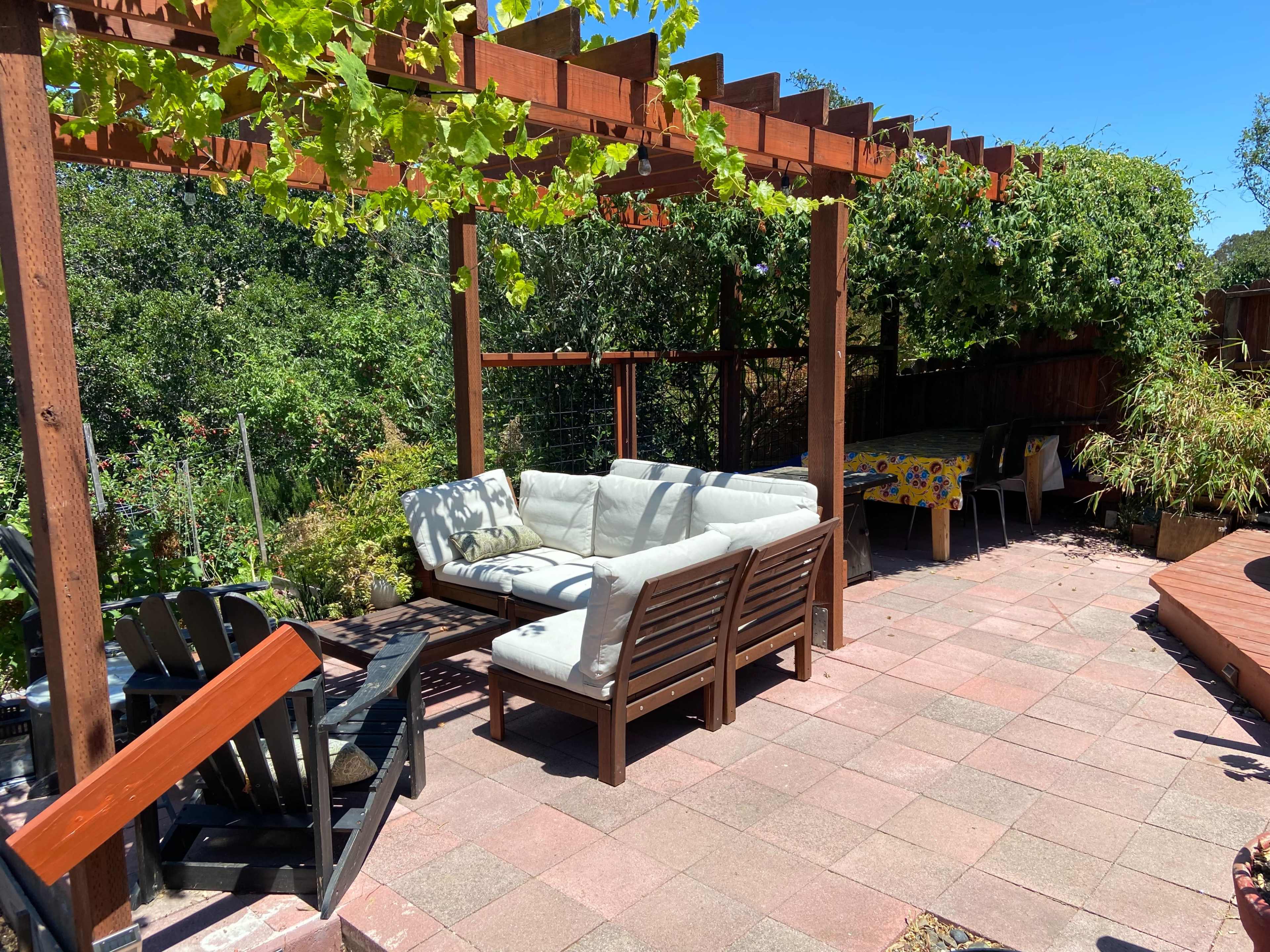 A patio area features a wooden pergola adorned with vines, a sectional sofa, and a dining table under bright sunlight.
