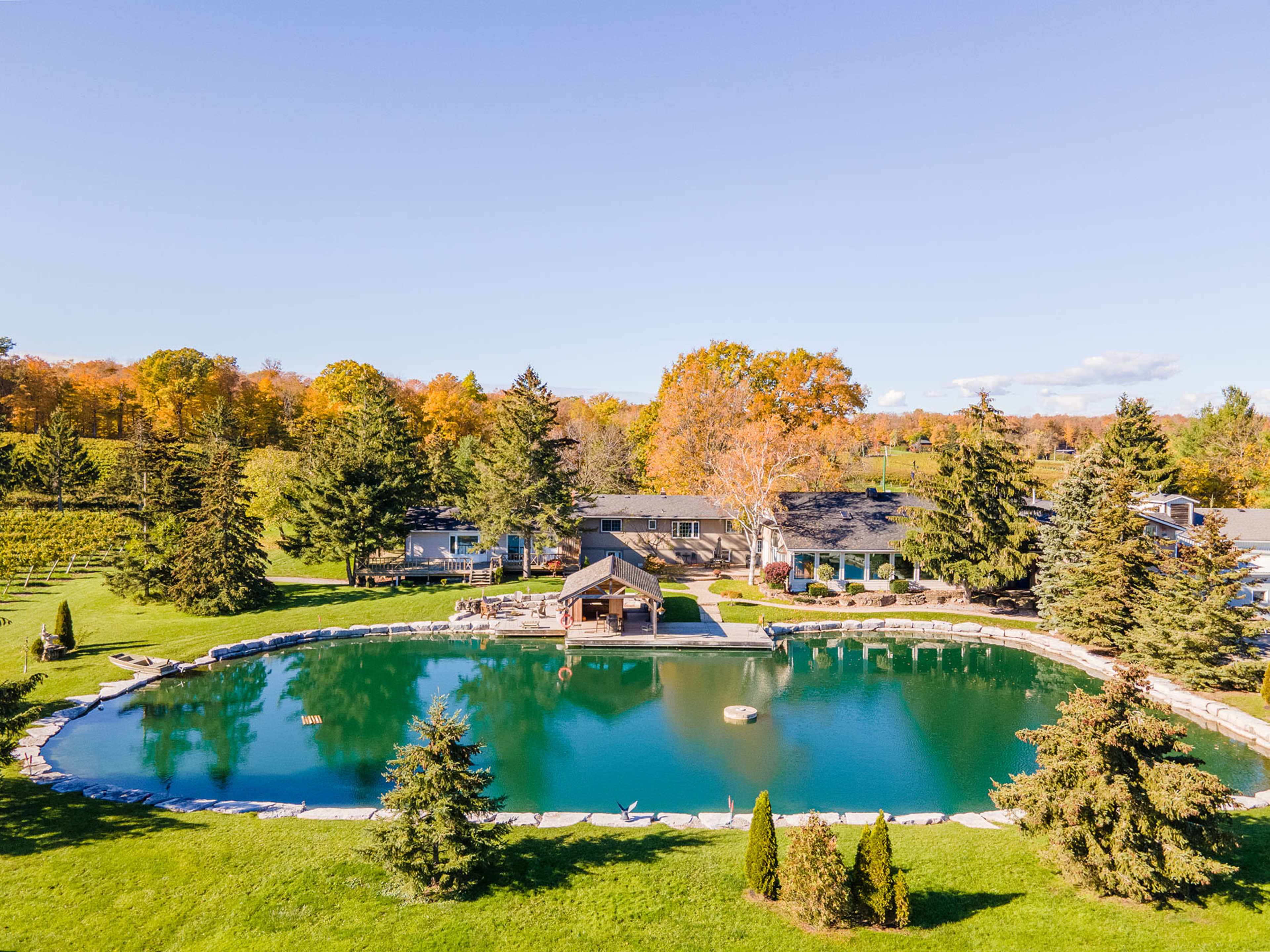 A large, serene pond surrounded by trees and a stone edge is located in front of a house and a gazebo.
