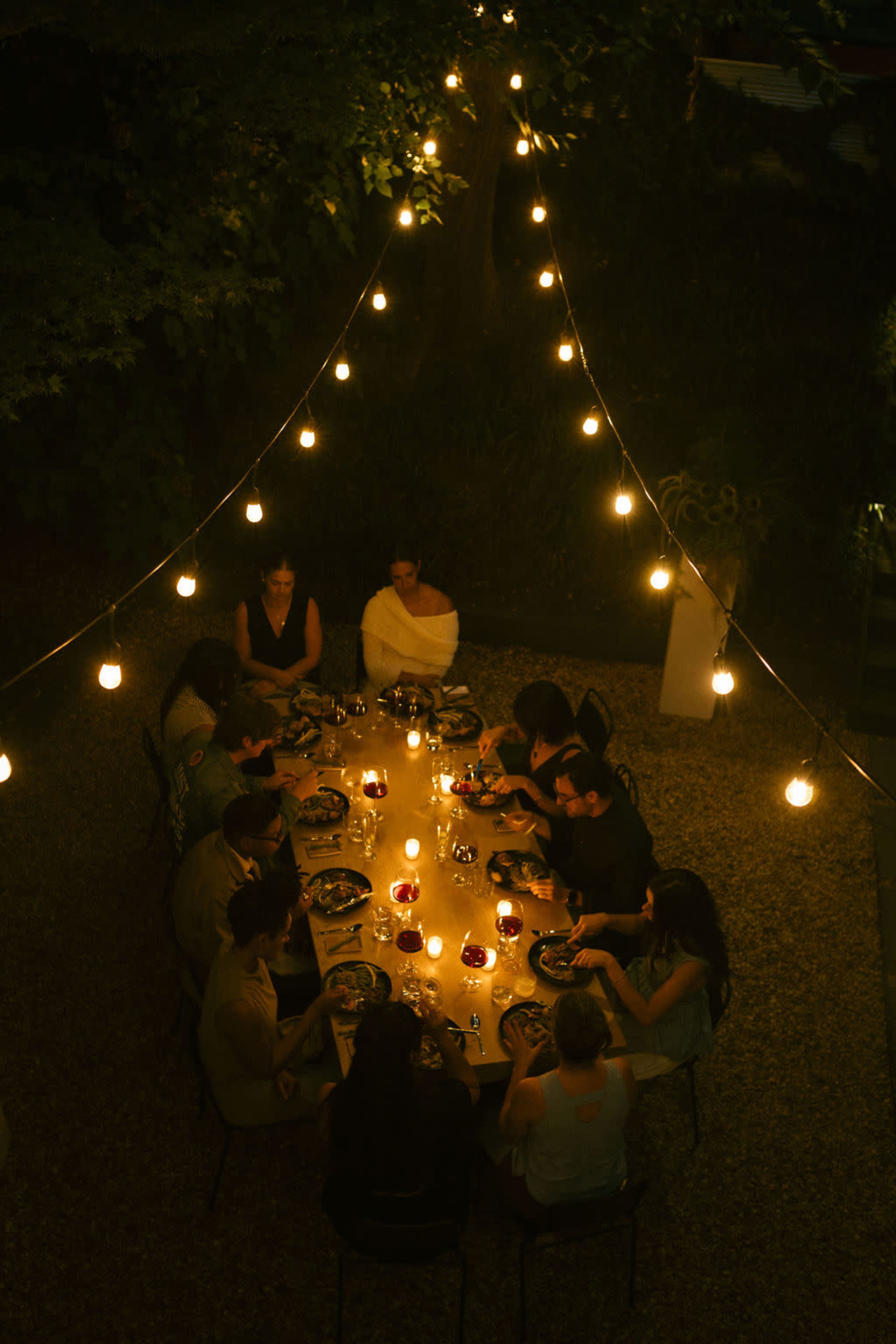 A long table set for dinner is surrounded by people dining under overhead string lights in an outdoor setting.