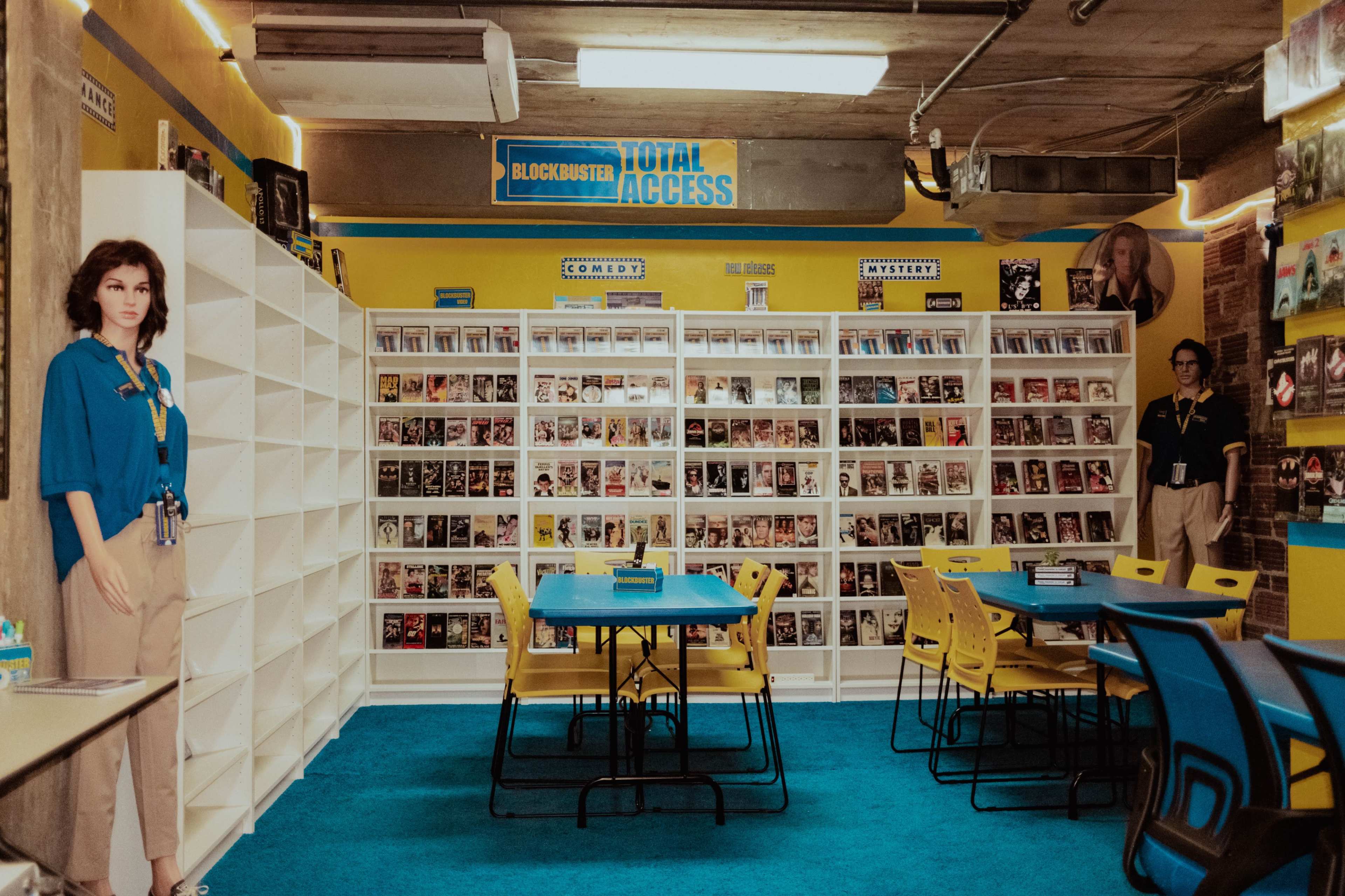 The image shows an interior of a Blockbuster store, featuring movie displays, rows of DVDs, and seating areas with bright blue carpet and colorful chairs.