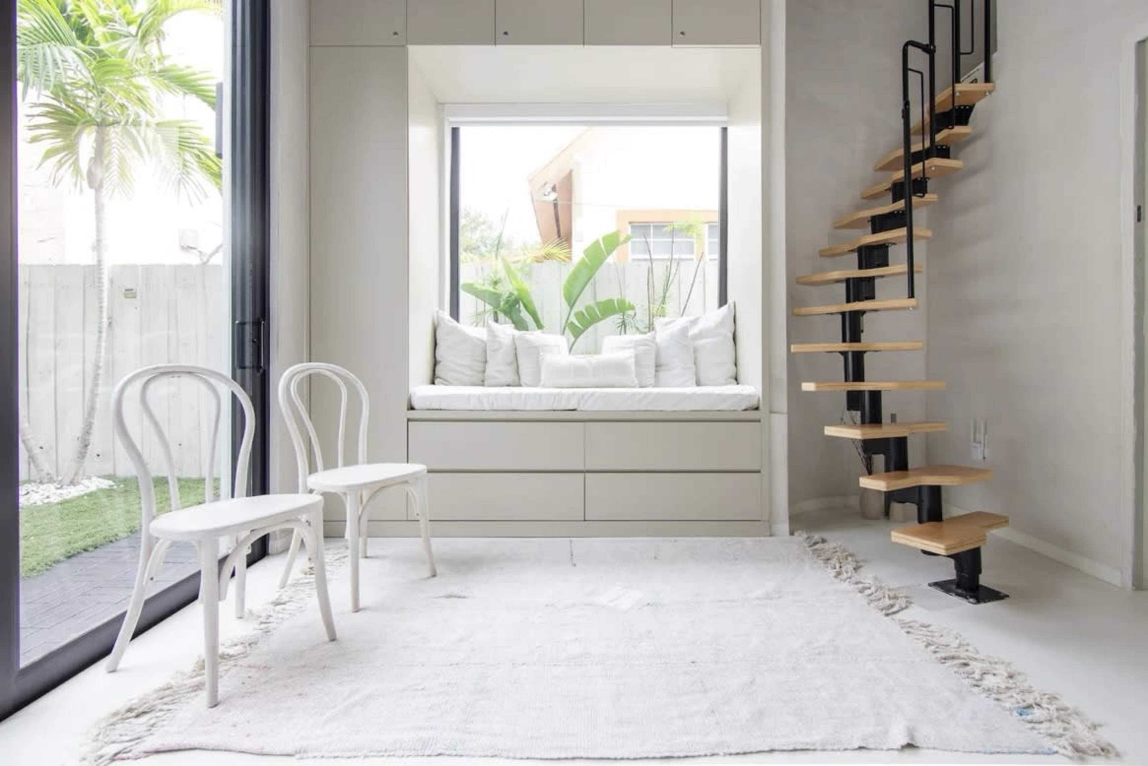 The image shows a bright interior space featuring a window seat with cushions, two white chairs, a wooden spiral staircase, and a light-colored rug on the floor.
