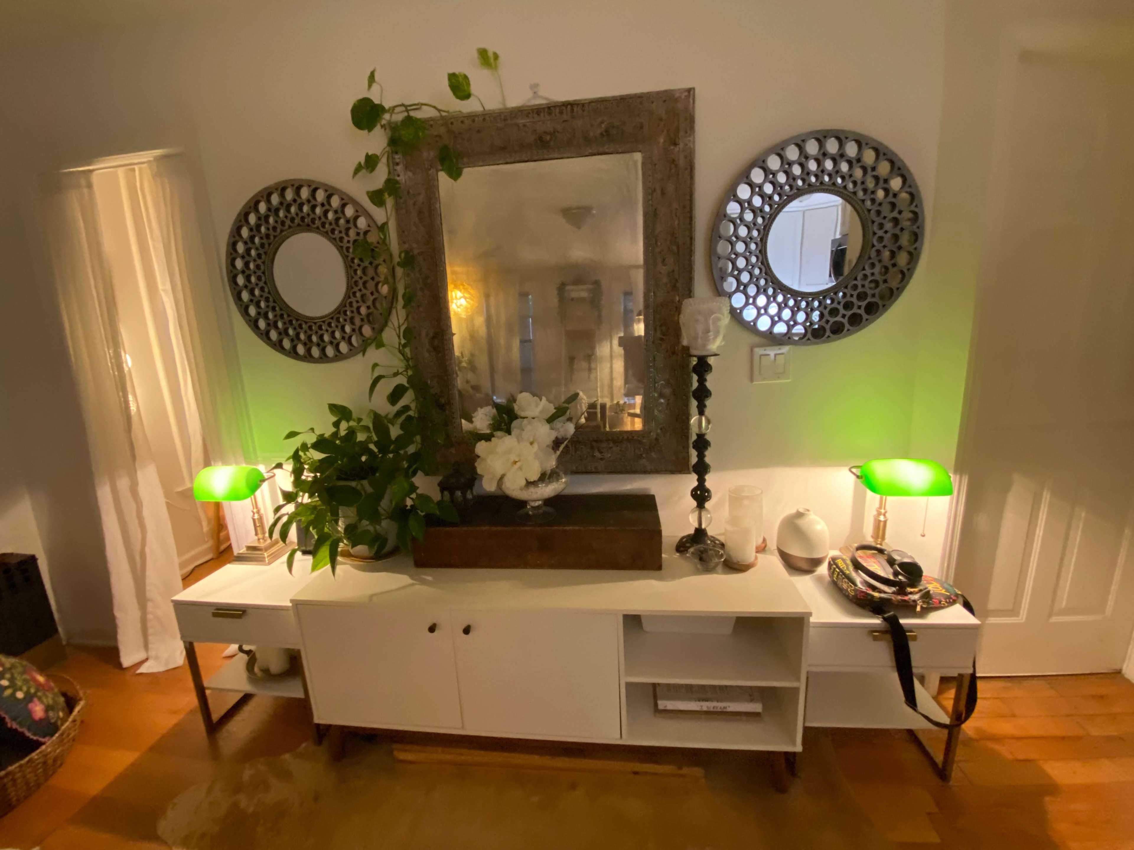 A console table with a large mirror and decorative mirrors, flanked by two green lamps and adorned with plants and flowers.