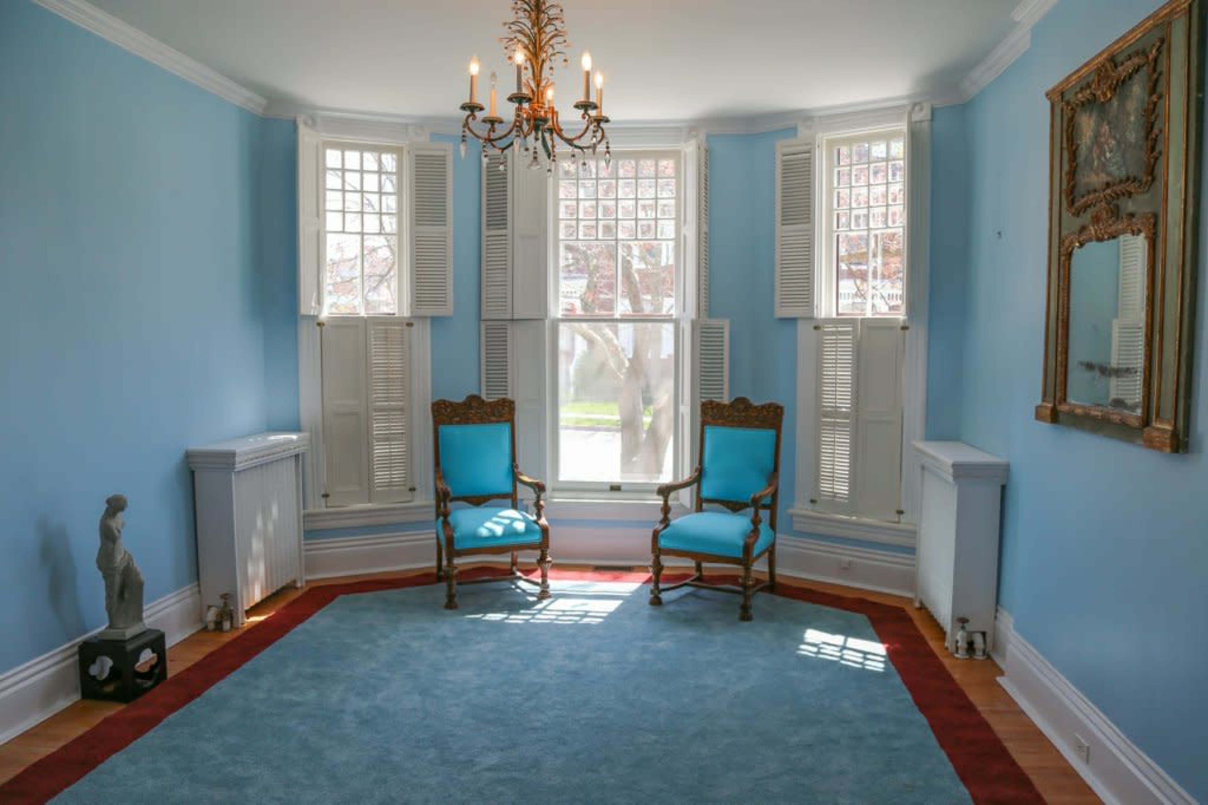 A room with light blue walls features two wooden chairs, a chandelier, and large windows with shutters.